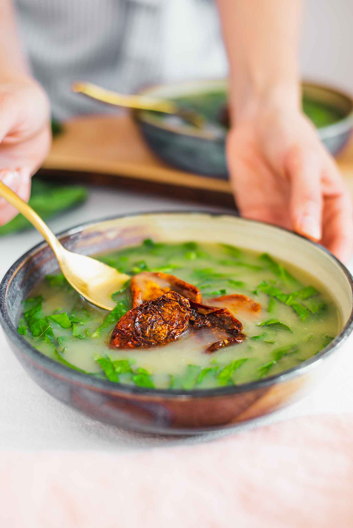Side view of a hand holding a spoon and dipping it into the vegan Portuguese collard soup. Collard greens swirl in the broth and smoky sun-dried tomatoes garnish the top.