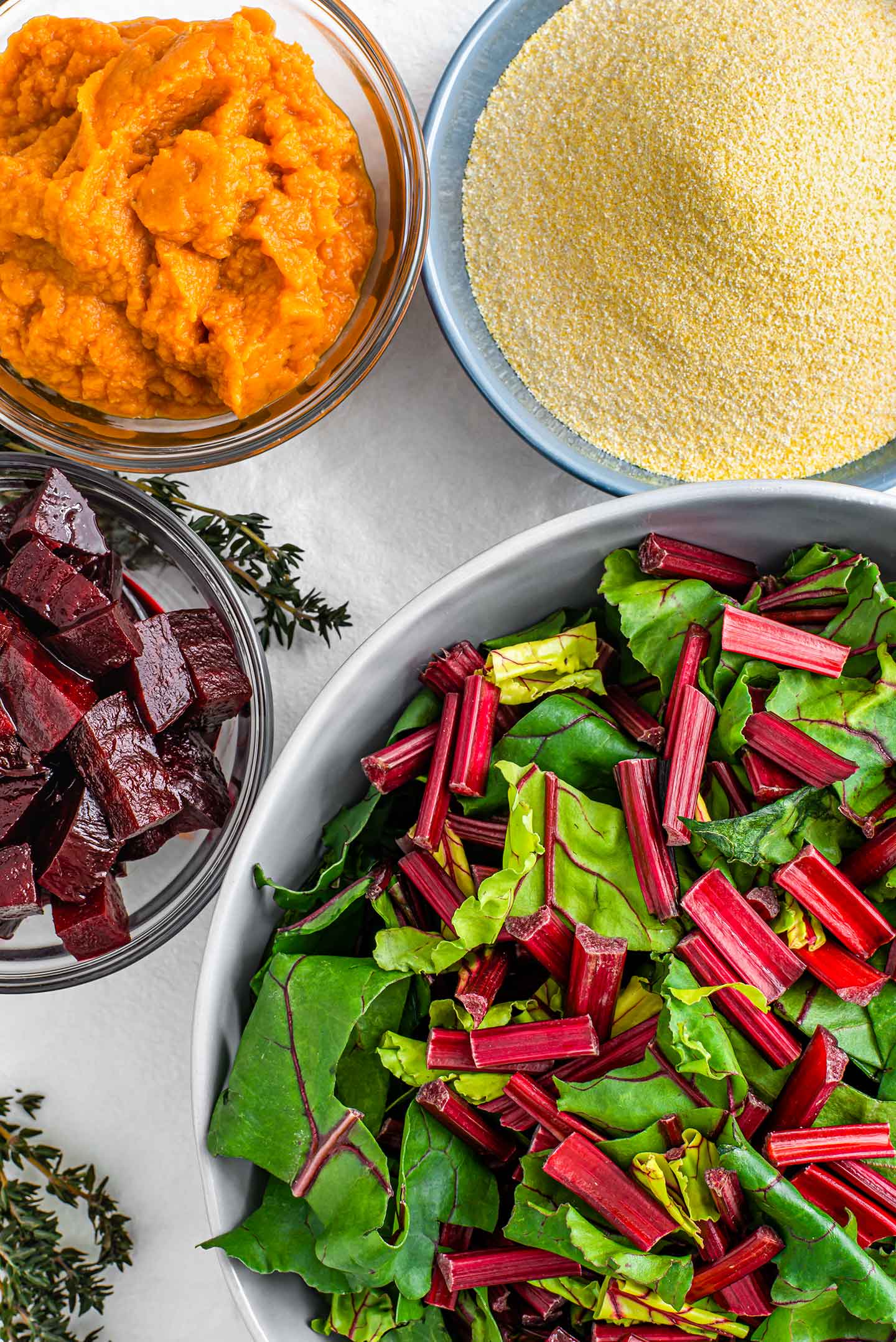 Top down view of ingredients for festive pumpkin beet polenta. Raw beet greens, roasted beets, pumpkin puree, and corneal fill separate dishes on a white tray.