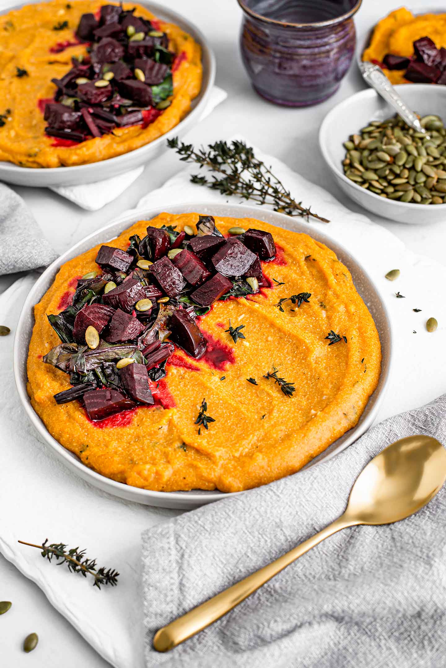 Top down view of two plates of festive pumpkin beet polenta. The orange coloured polenta is topped with the deep purple beets, and sprinkled with pumpkin seeds and thyme.