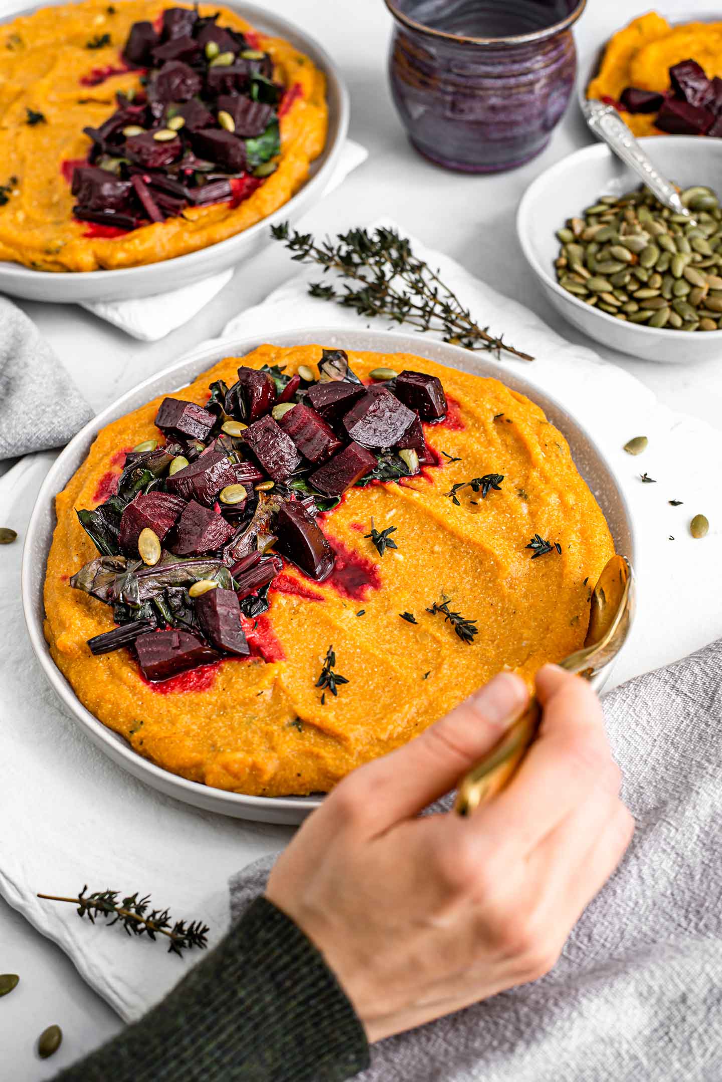 Side view of a hand with a spoon, scooping into a plate of festive pumpkin beet polenta. Another dish stands in the background, and pumpkin seeds fill a small bowl.