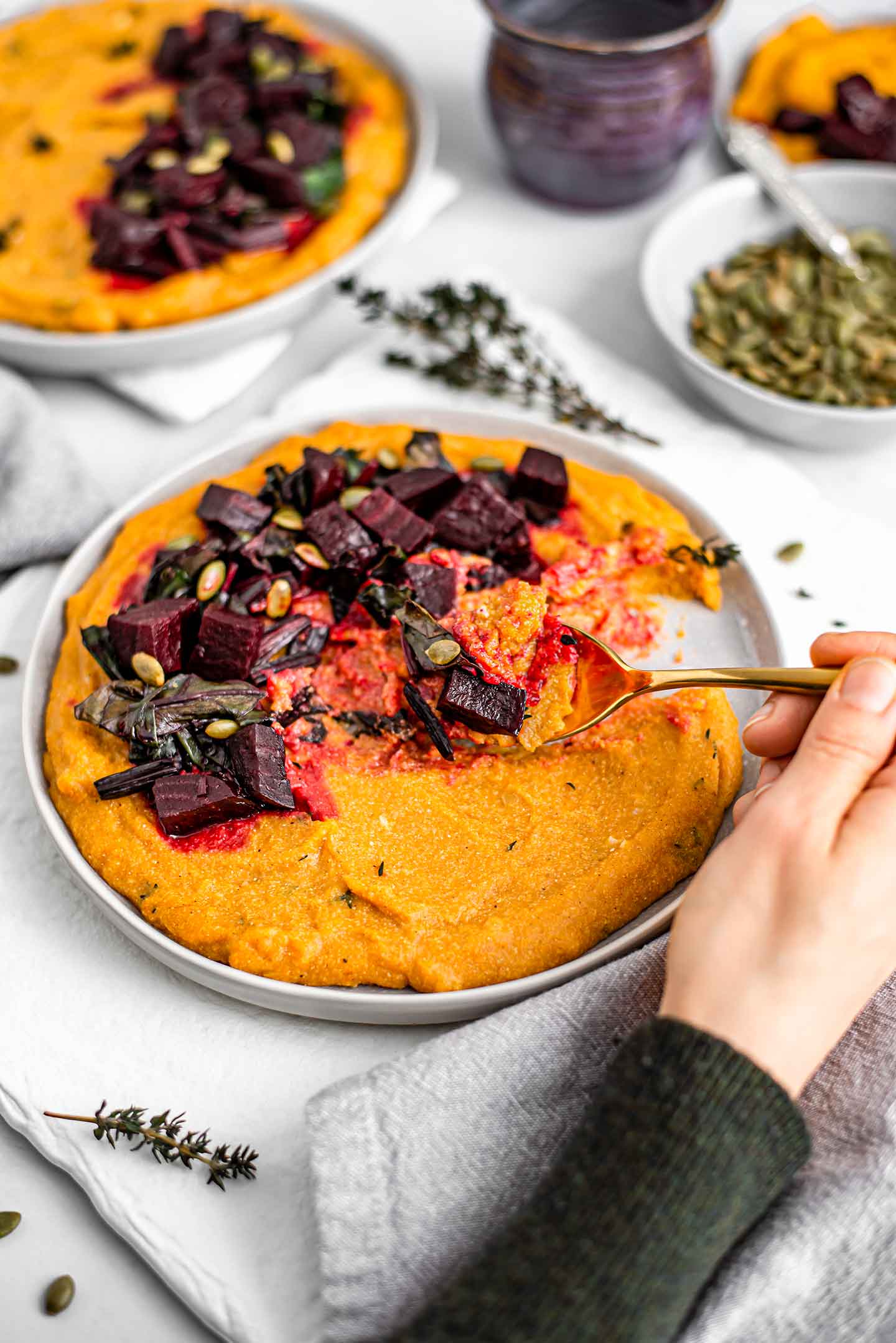 Top down view of a hand holding a spoon that is scooping up the polenta. The orange pumpkin flavoured polenta swirls with the purple colour from the beets.