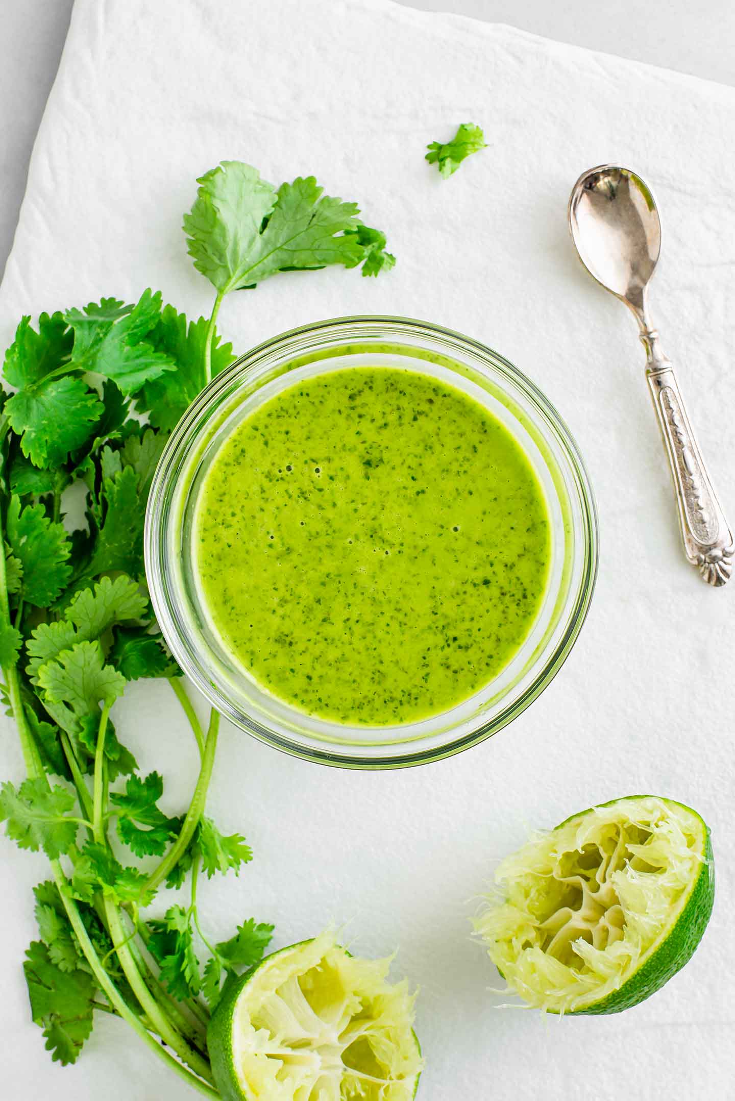 Top down view of bright green cilantro lime vinaigrette filling a small dish. A spoon lays beside the dish, as do fresh cilantro leaves, and a squeezed lime.