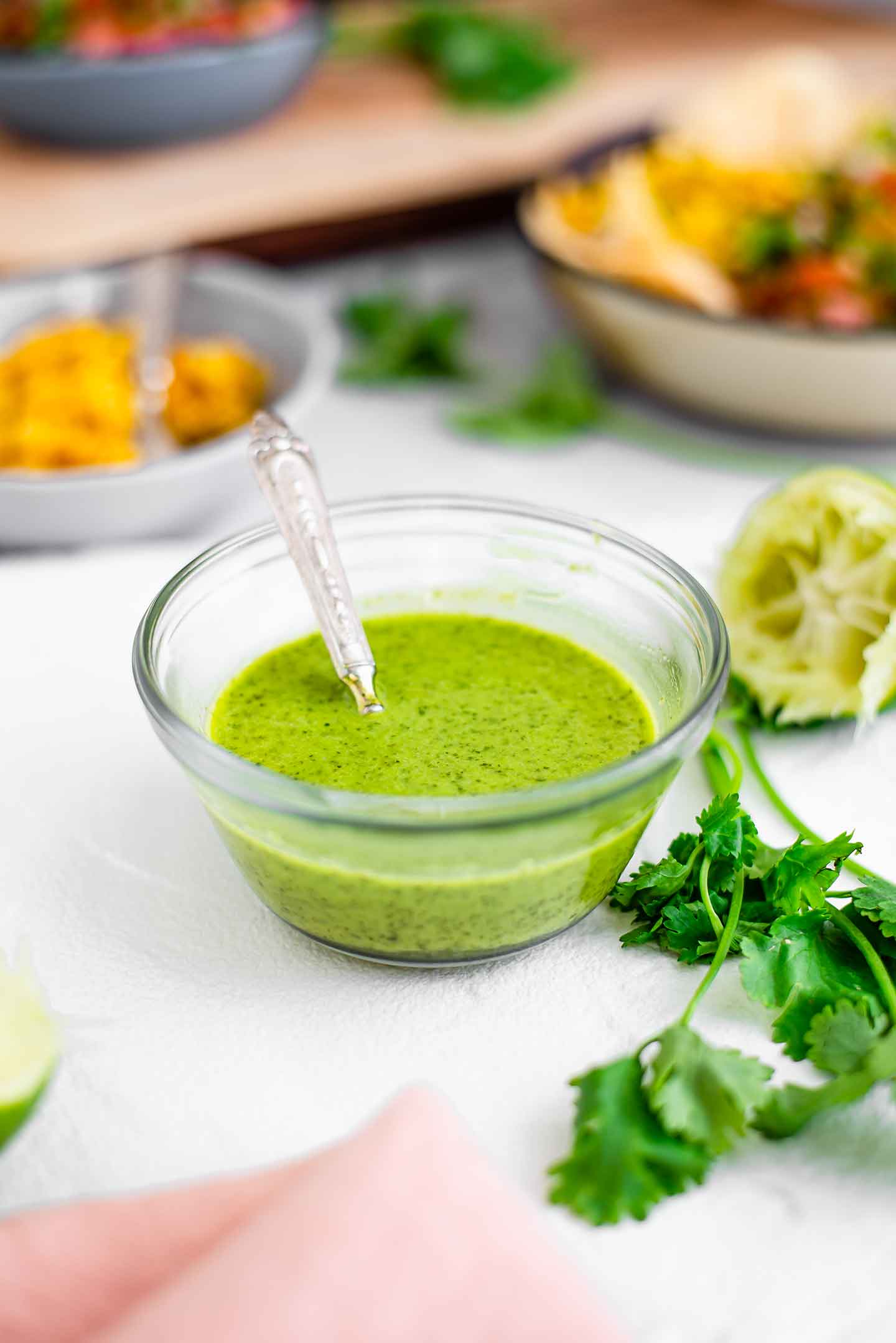 Side view of cilantro lime vinaigrette in a small dish with a spoon. Cilantro leaves and squeezed lime surround the bowl. Salad ingredients can be seen in the background.