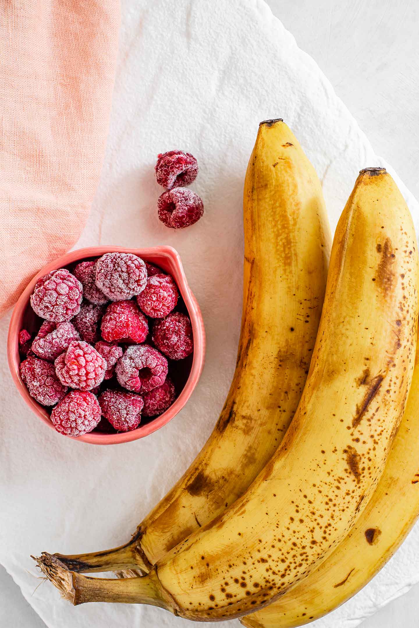 Top down view of three yellow, spotted bananas and frozen raspberries on a white tray.