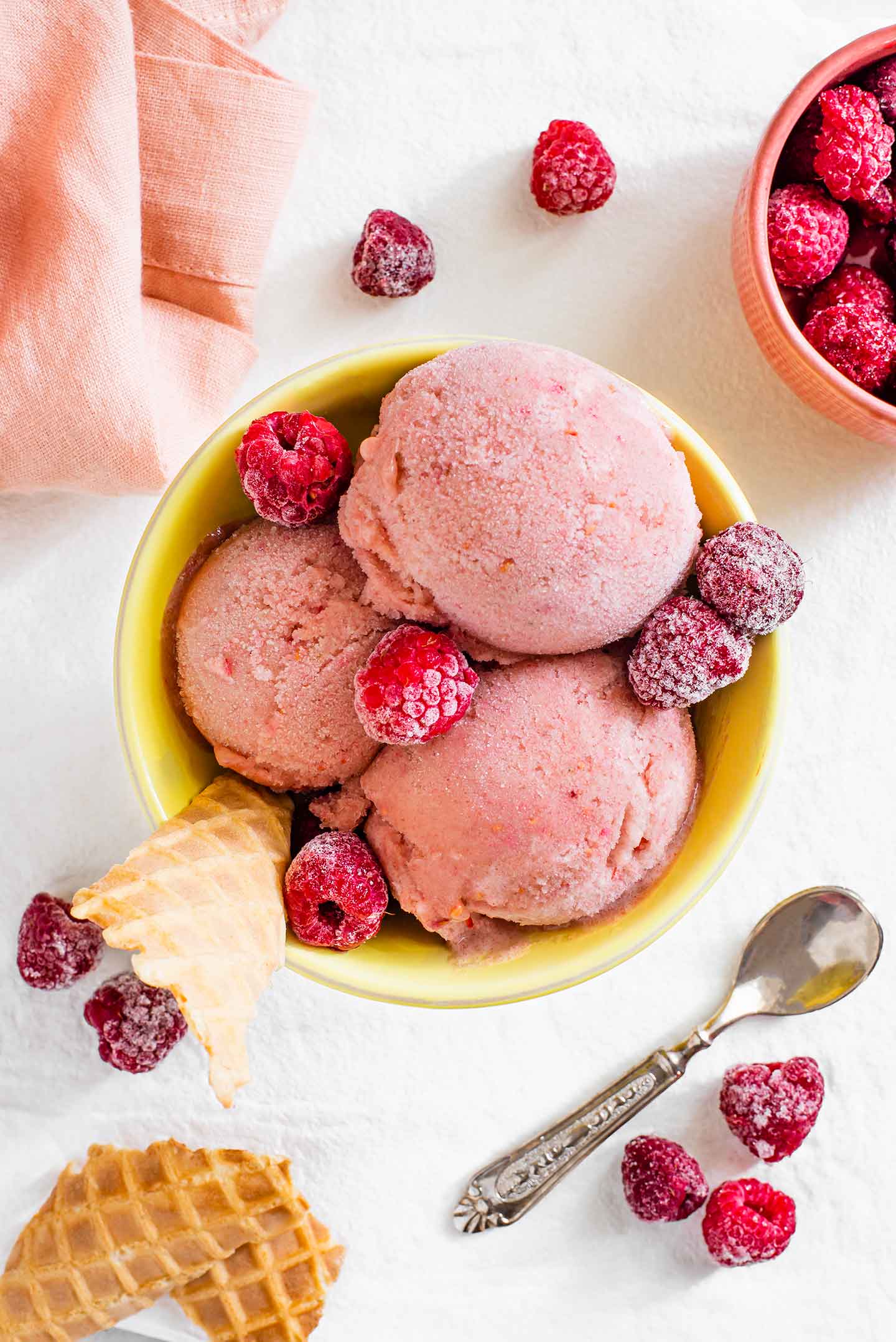 Top down view of three pink scoops of raspberry nice cream in a yellow sorbet cup with frozen raspberries and pieces of waffle cone scattered around.