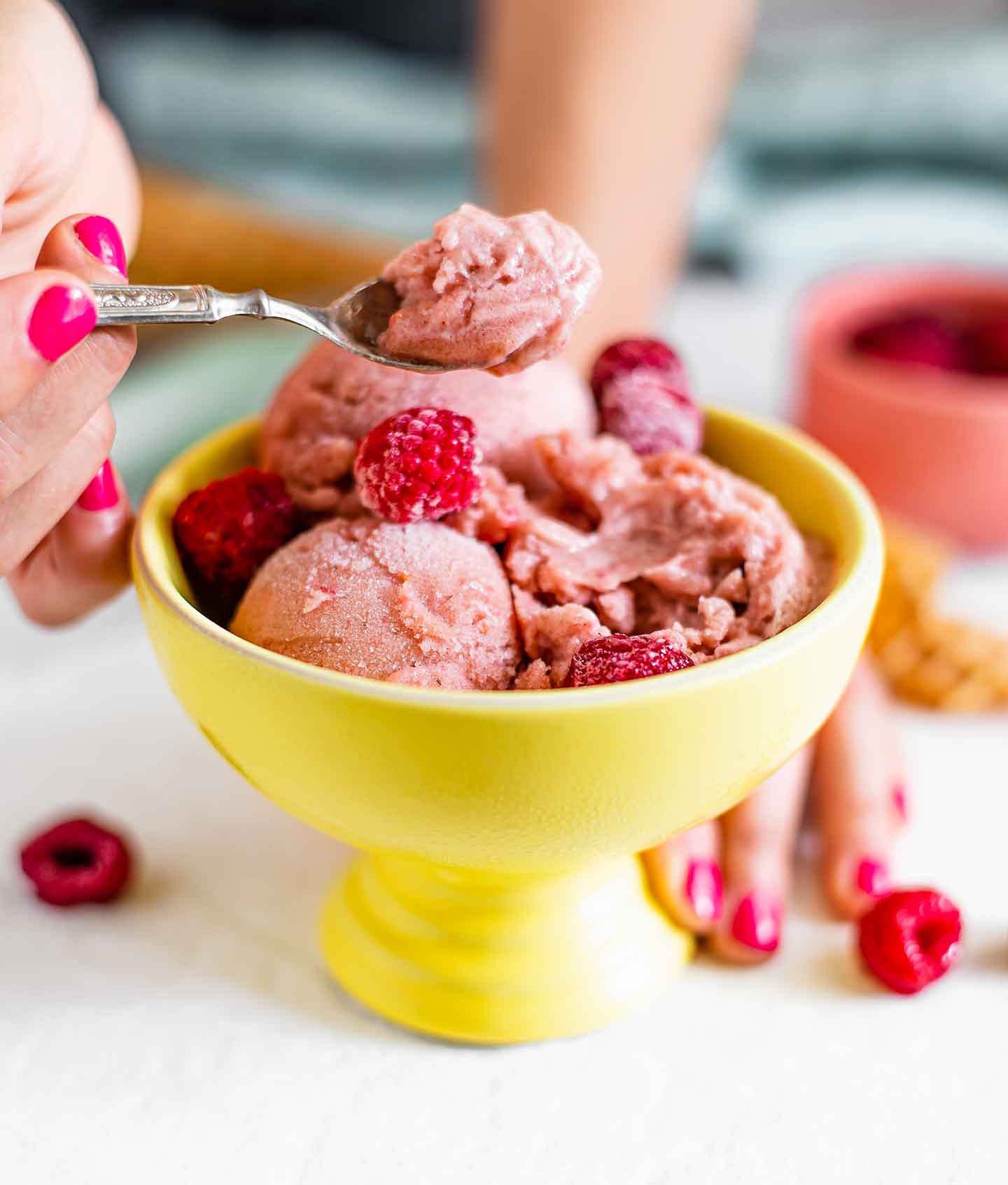 Side view of hand scooping raspberry nice cream on a small spoon from a sorbet cup. The nice cream is bright pink and creamy.