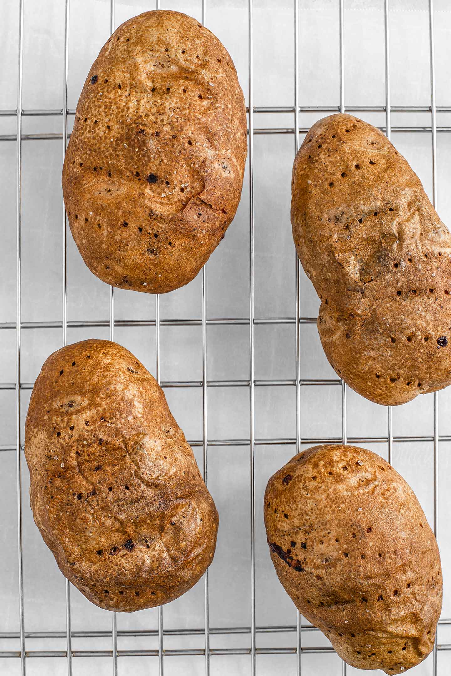 Top down view of four baked potatoes on a wire cooling rack. The skin is wrinkly, crispy, and the small holes are visible from being pierced with a fork before baking.