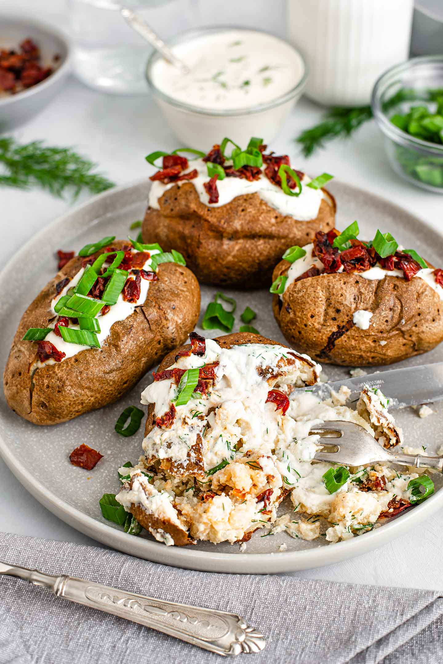 Three loaded potatoes and one smashed potato are on a plate. A fork rests in the smashed potato. Soft potato mixes with sour cream, sun-dried tomato bits, and green onion.