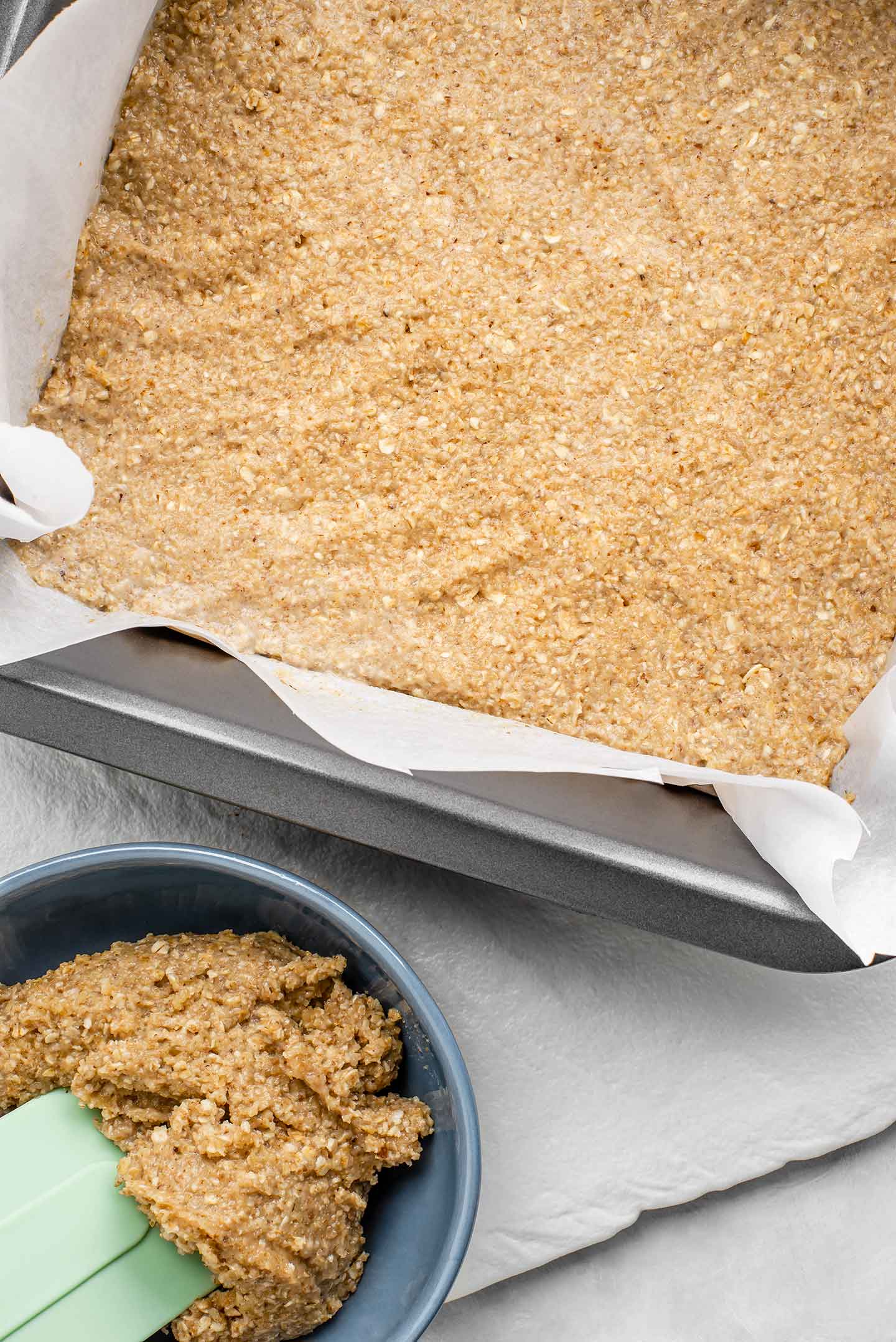 Top down view of an oat, walnut crust packed into a baking tin lined with parchment paper. A scoop of dough is reserved in a small bowl to the side.