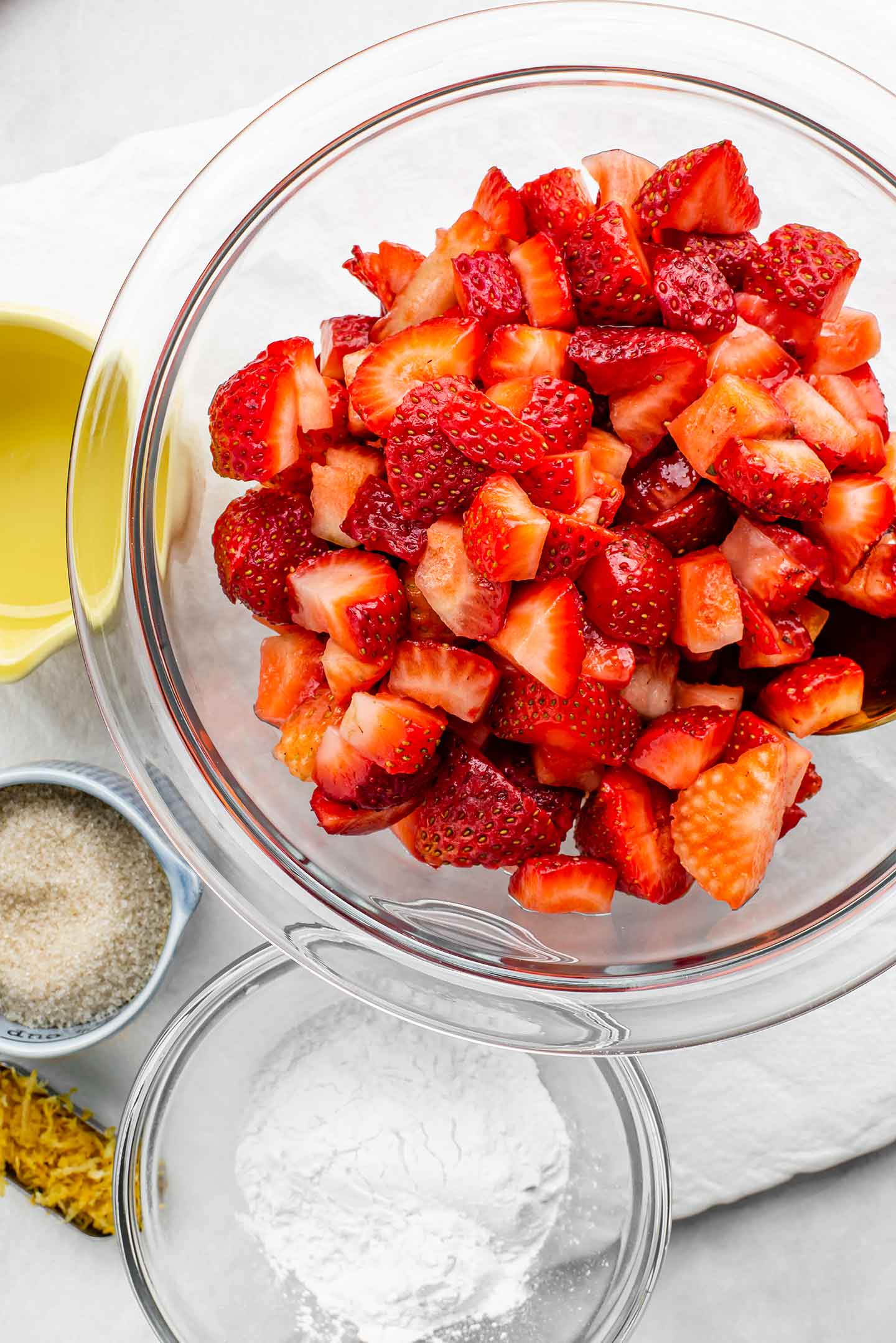 Top down view of diced strawberries in a glass bowl with lemon juice, cane sugar, lemon zest and arrowroot starch in separate dishes around the berries.
