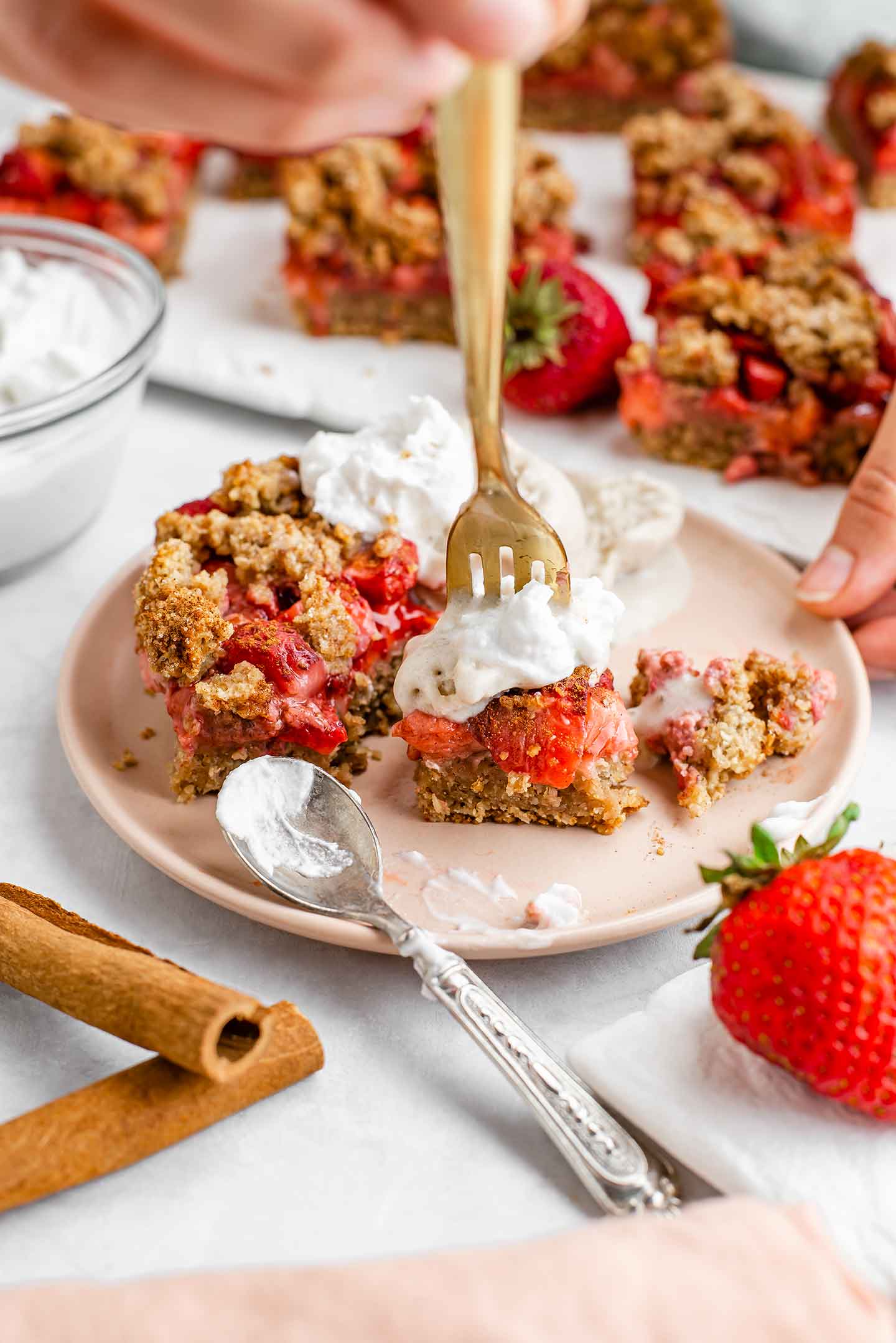 Side view of a strawberry crumble topped with ice cream and whipped cream on a fork. Strawberry bars fill a tray in the background.