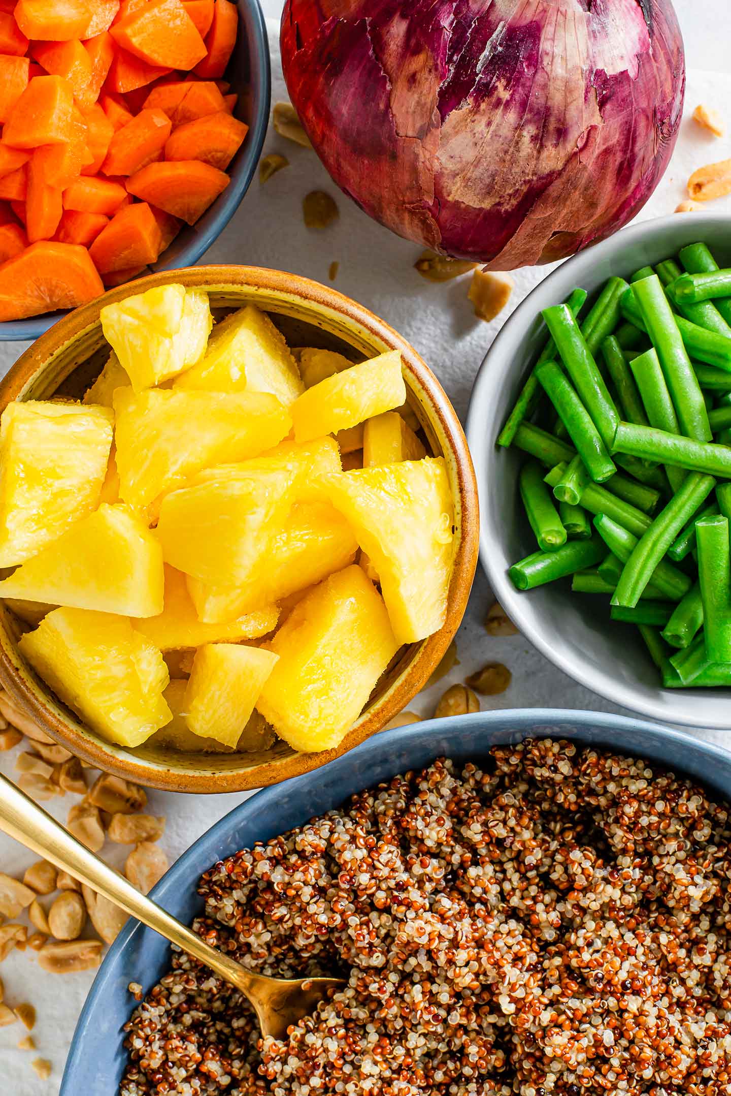 Top down view of pineapple, carrots, green beans, a red onion and a bowl of quinoa displayed on a white tray sprinkled with roasted peanuts.