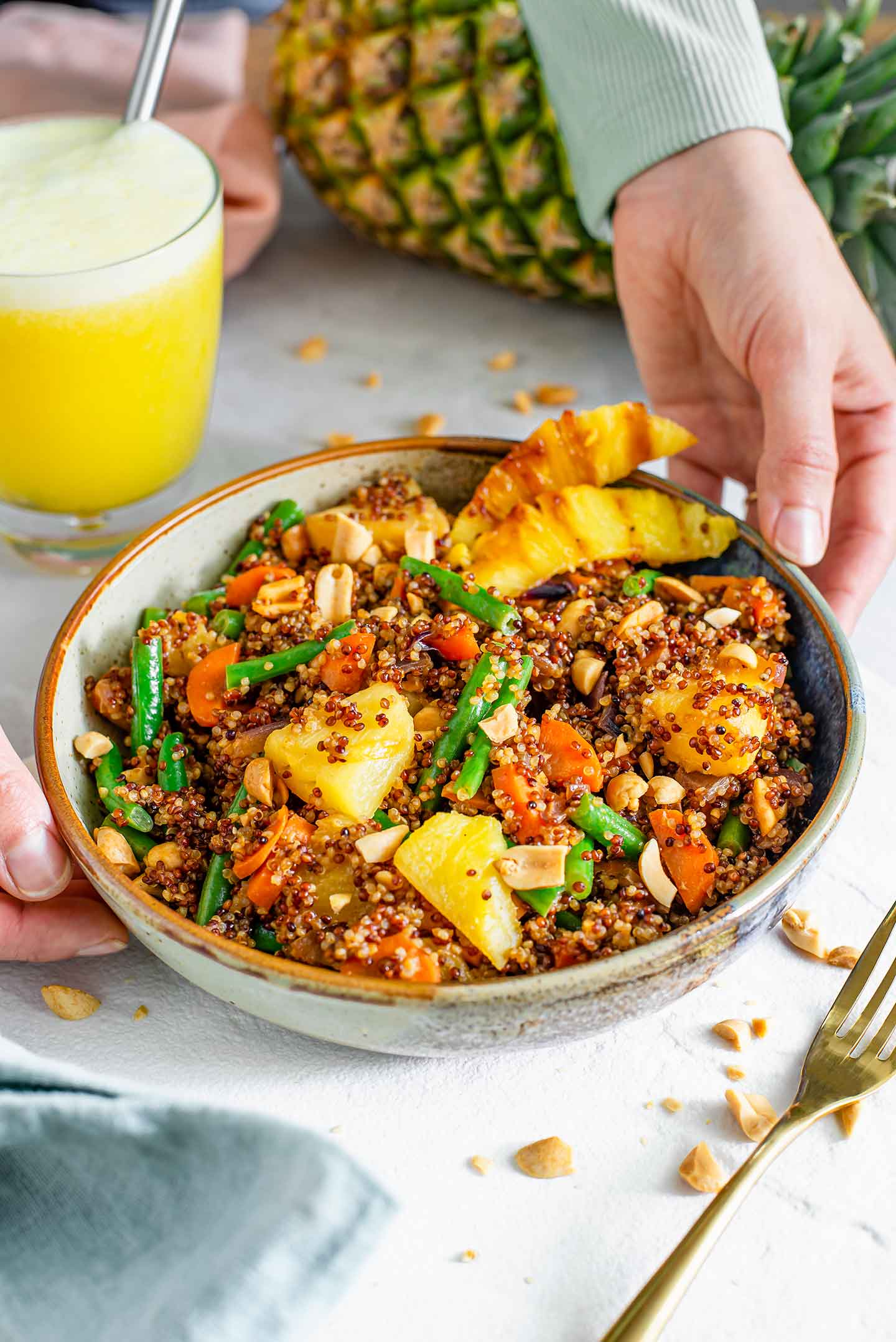 Side view of hands placing a shallow bowl of pineapple quinoa stir-fry on a white tray. Pieces of grilled and caramelized pineapple garnish the side of the dish.