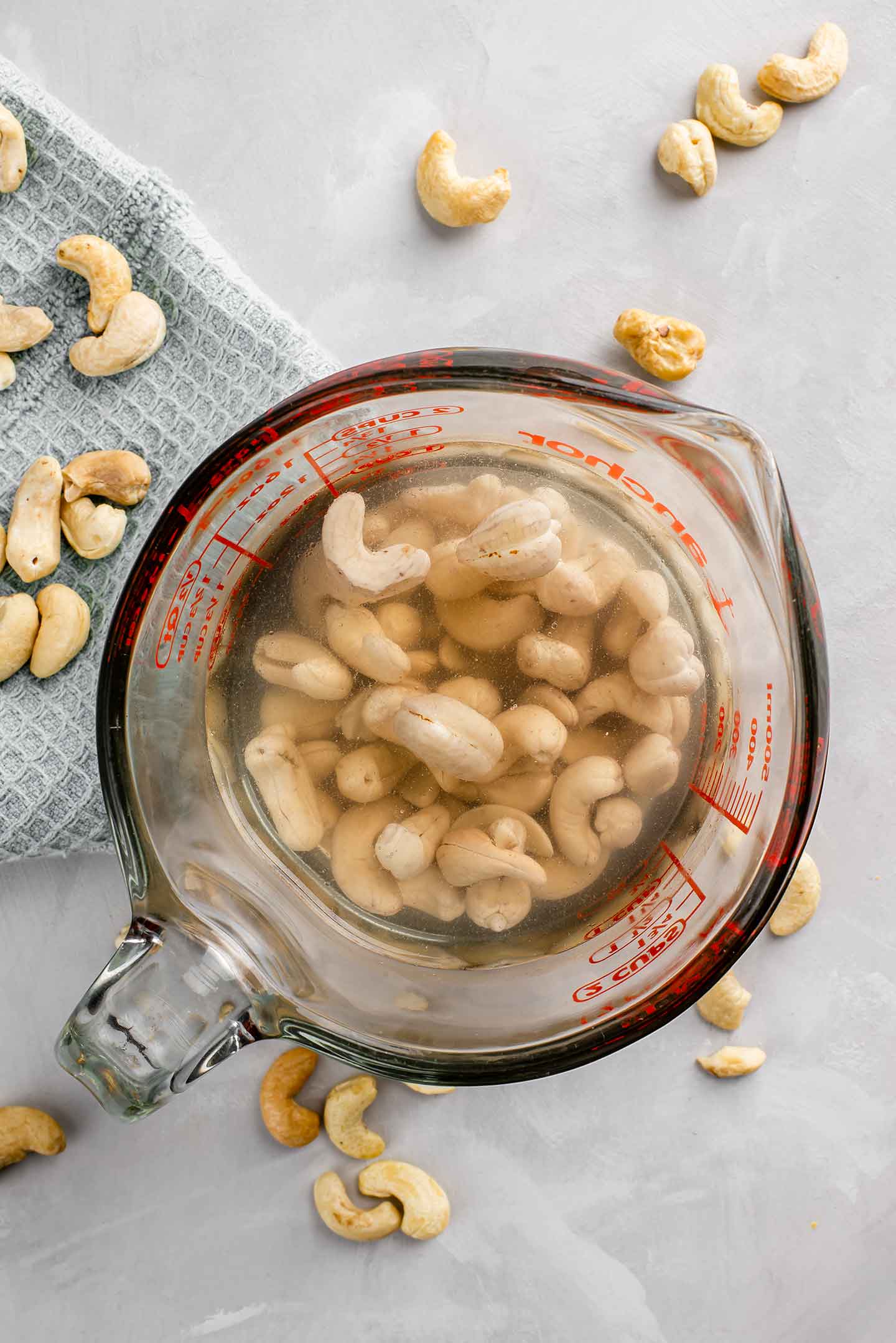 Top down view of cashews soaking in boiling water in a glass measuring cup with raw cashews scattered around.