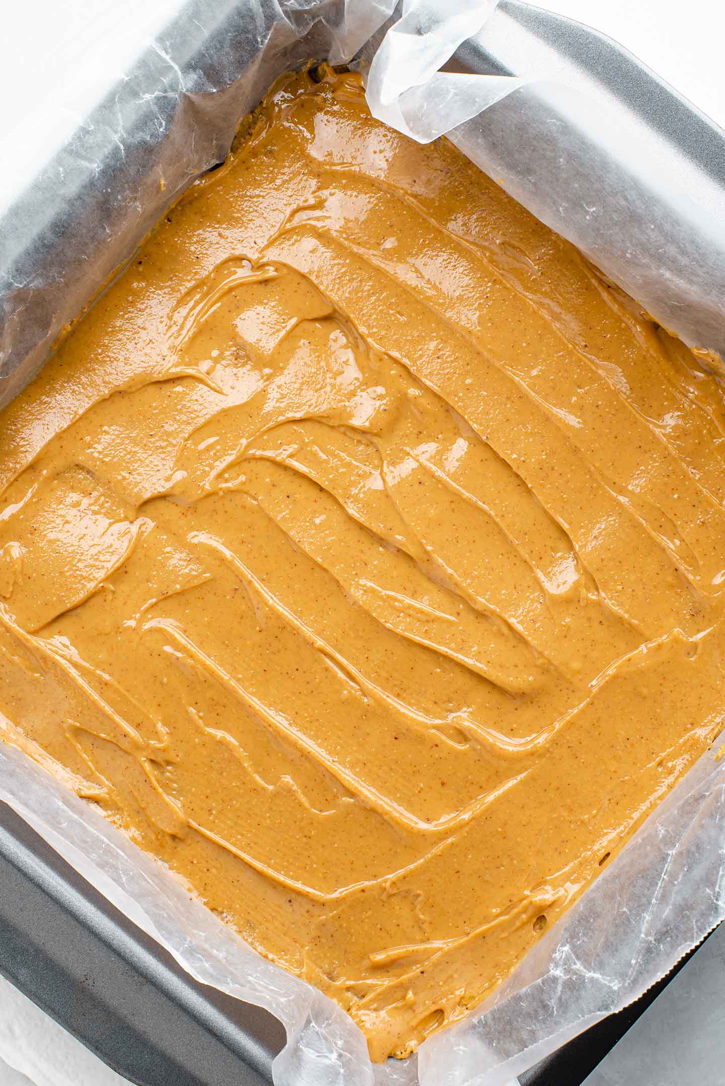 Top down view of peanut butter in a smooth layer on top of the base. The mixture is still in a wax paper lined baking tray.