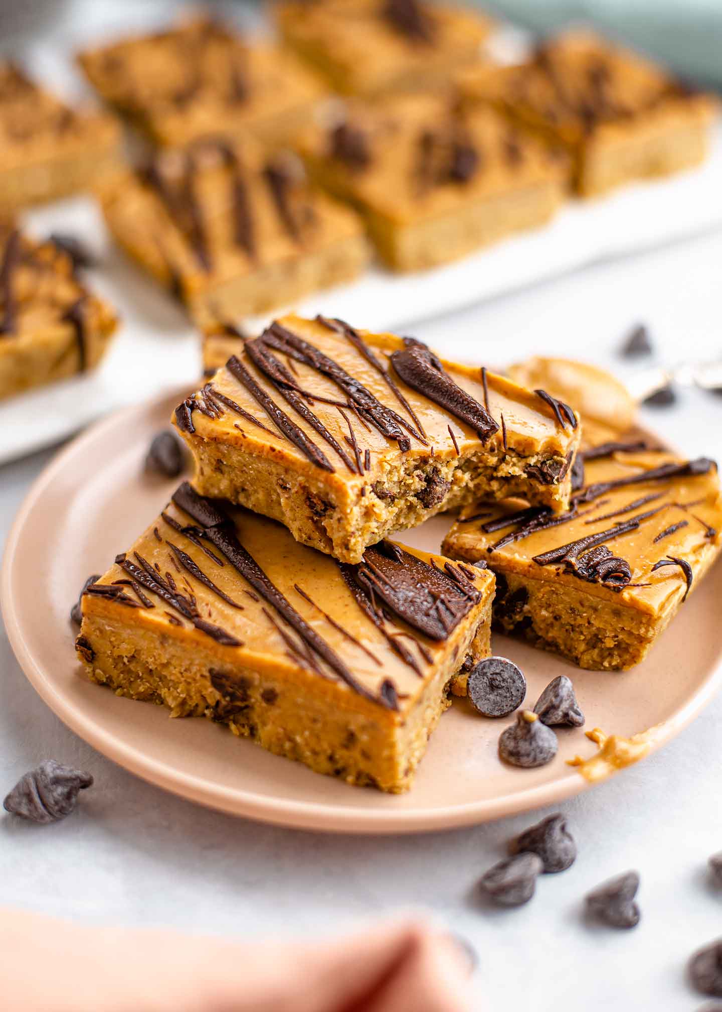A bite is taken out of a protein cookie dough bar speckled with chocolate chips, topped with a layer of peanut butter, and drizzled with chocolate. The bar rests on a stack of other bars displayed on a plate.