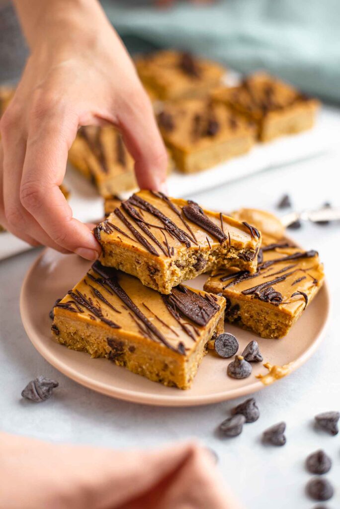 A bite is taken out of a chickpea cookie dough bar. A hand places the bar on top of a stack of other bars. A tray of bars is visible in the background.