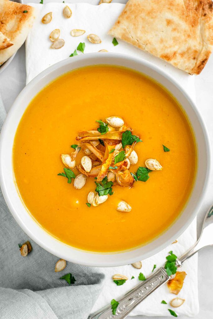 Top down view of curried butternut squash soup garnished with squash seeds, roasted squash skin, and fresh parsley. Naan bread and a spoon rest beside the soup.