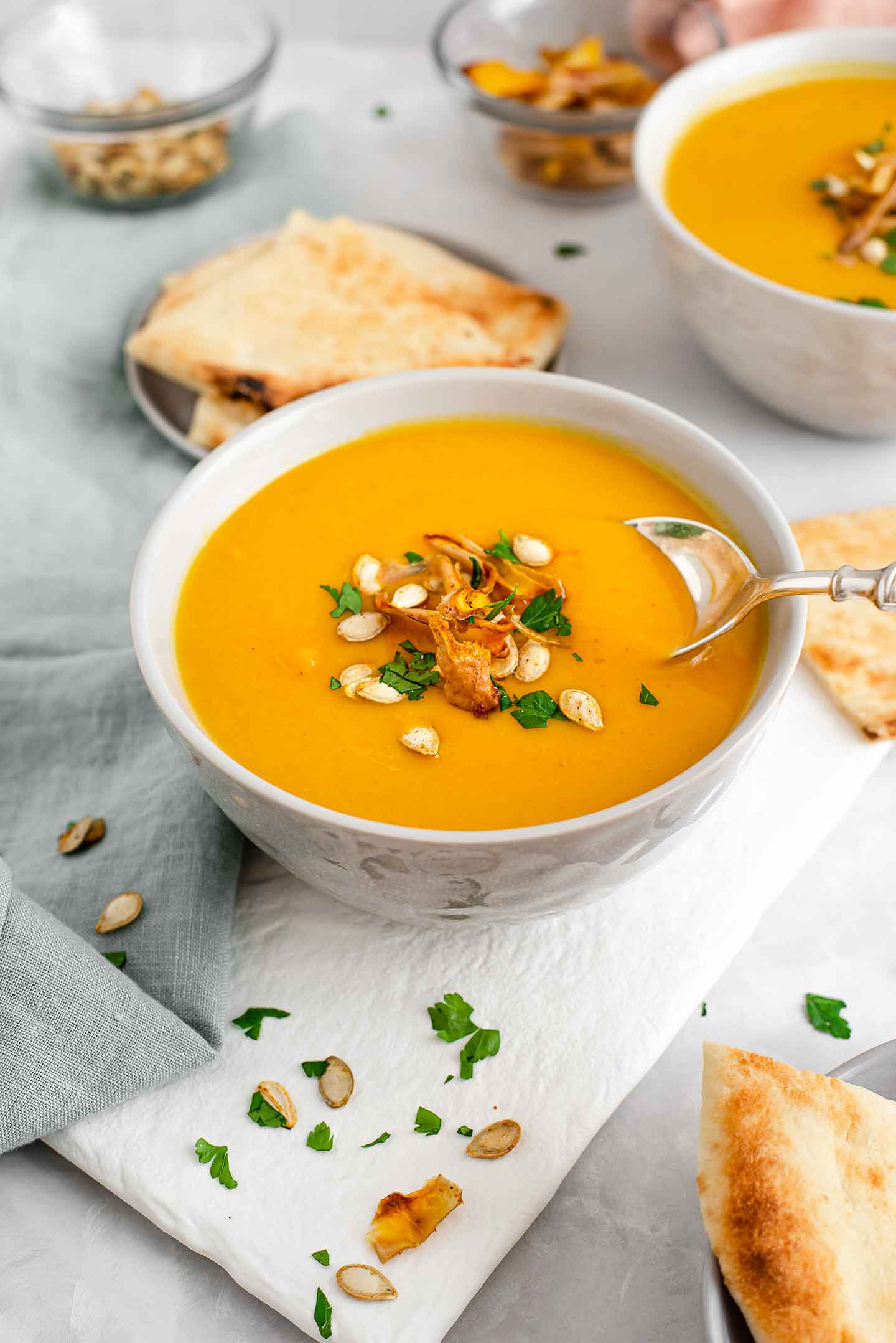 Side view of a spoon scooping curried butternut squash soup. The soup is a vibrant yellow/orange colour and topped with roasted squash seeds, squash skin chips, and fresh parsley. Naan bread is served on the side.