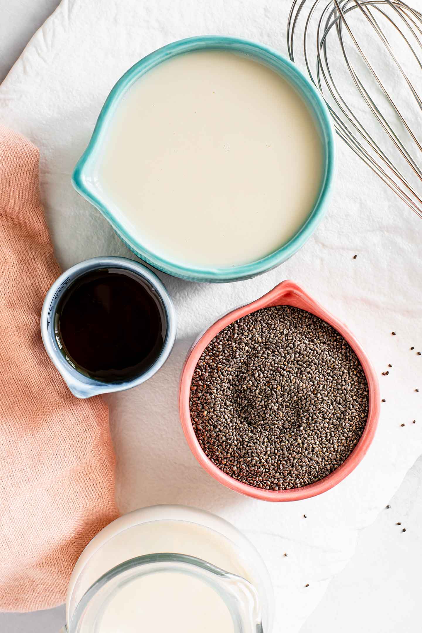 Top down view of black chia seeds in a small dish; with a whisk, a measuring cup of milk, and a small dish of maple syrup on top a white tray.