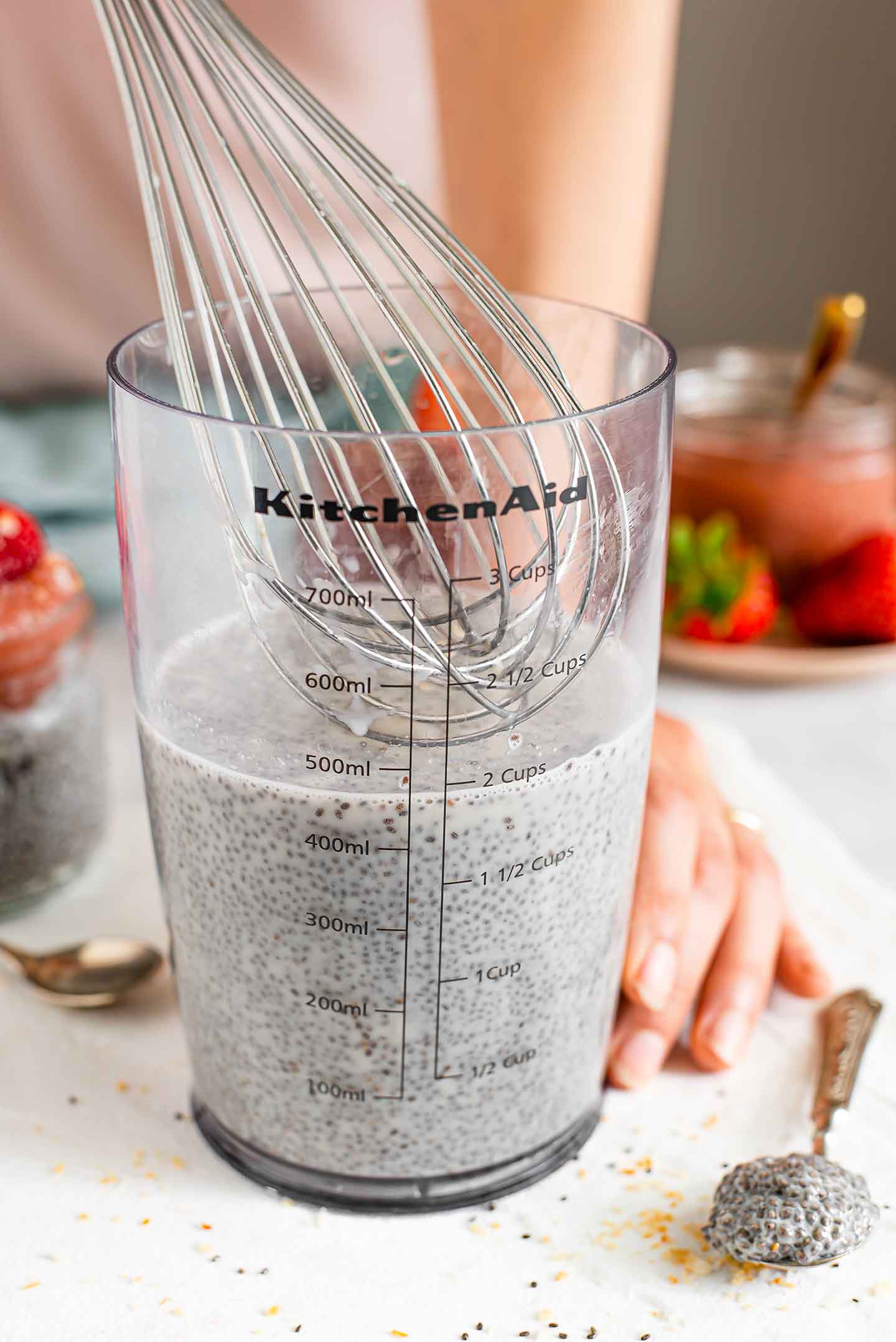 Side view of the seeds and milk being whisked in a clear container. Strawberries and jam are visible in the background.