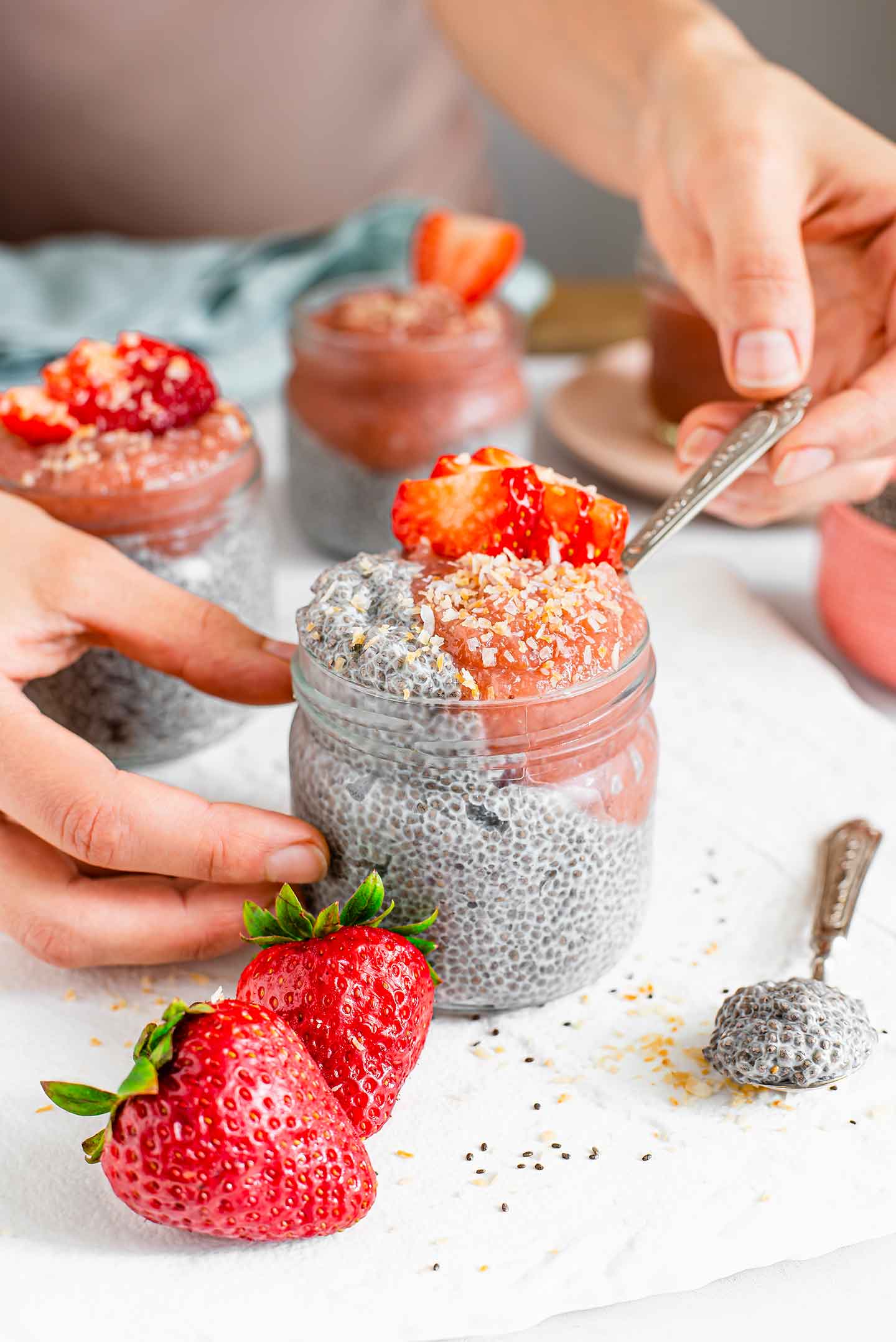 Side view of a hand dipping a spoon into a small glass jar of creamy chia pudding. The chia seeds are visible in the white pudding, pink jam fills a corner of the jar and the pudding is topped with sliced strawberry and toasted coconut.