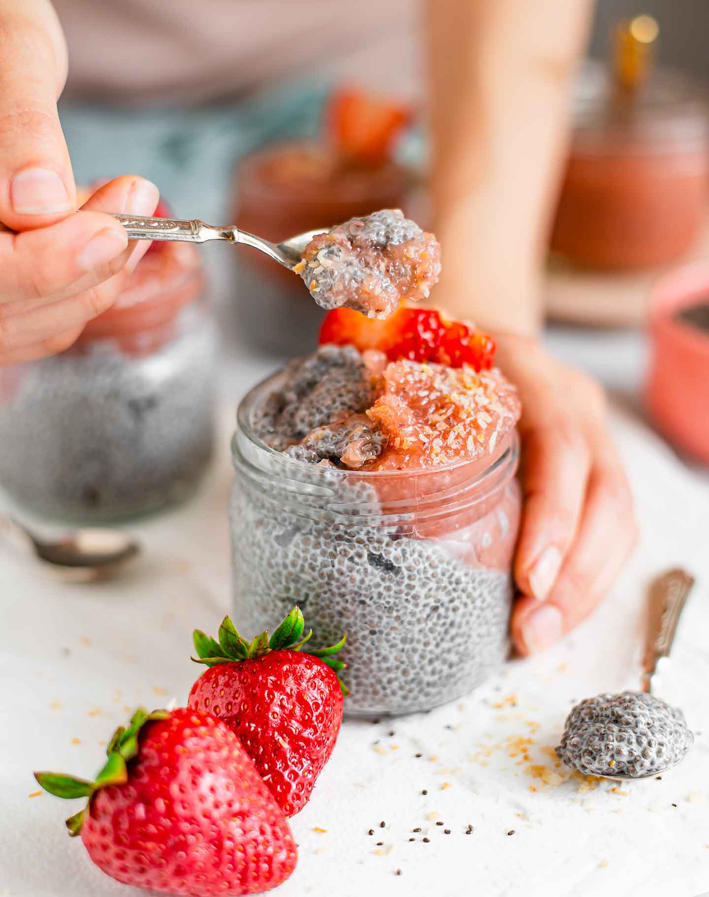 Side view of a hand scooping thick chia pudding from a jar. The pudding is topped with pink jam, fresh strawberries, and toasted coconut flakes.