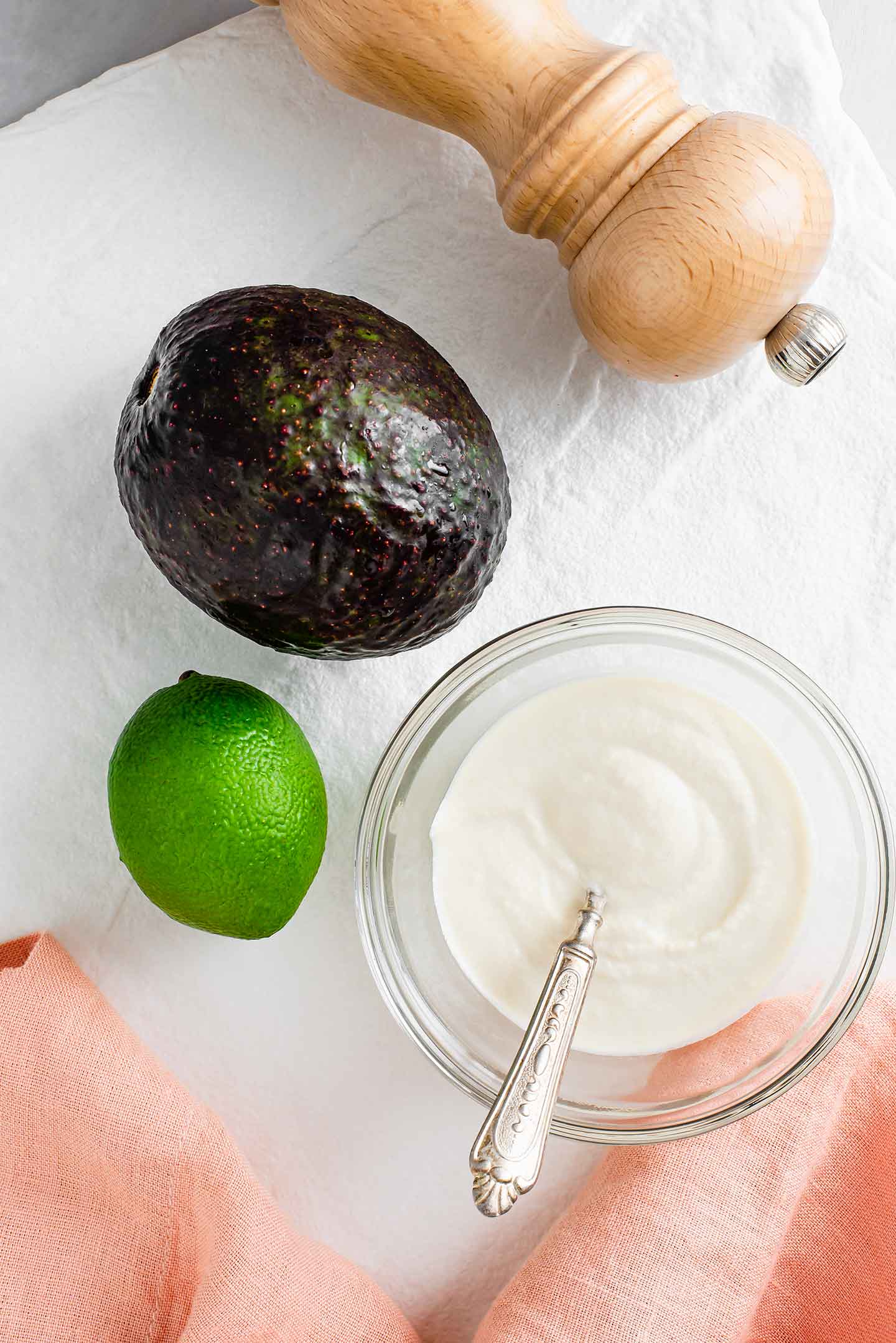 Top down view of an avocado, a lime, a small dish of sour cream, and a salt grinder displayed on a white tray.