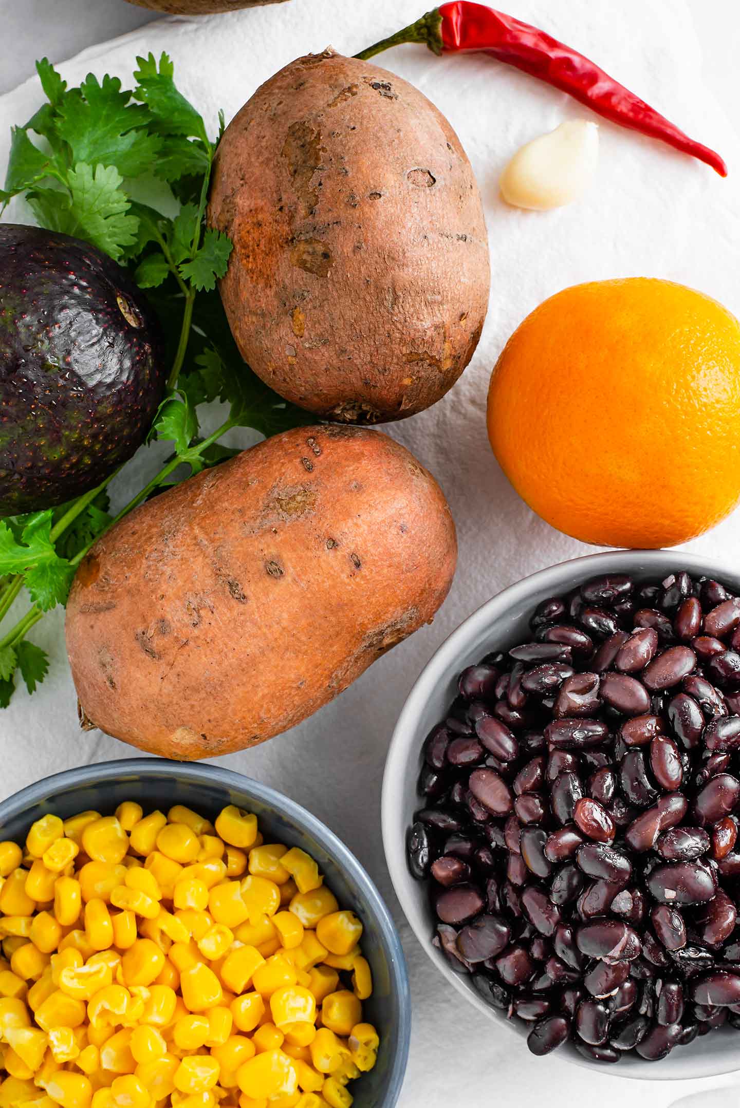 Top down view of ingredients on a white tray. Sweet potatoes, corn, black beans, an avocado, an orange, cilantro, a hot chilli pepper, and a clove of garlic are pictured.