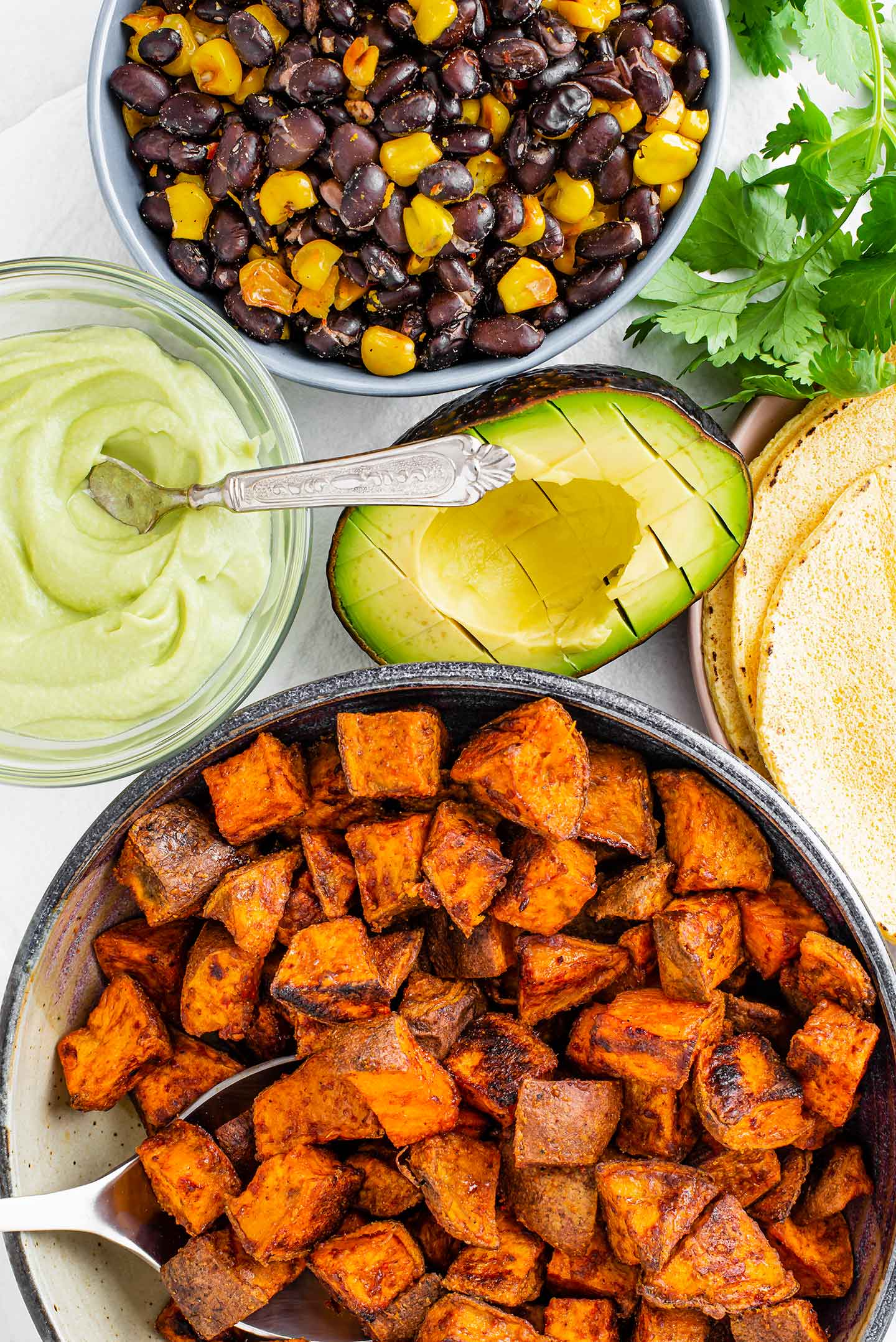 Top down view of roasted sweet potato, charred corn and black beans, a scored avocado half, corn tortillas, and a small dish of avocado lime crema. 