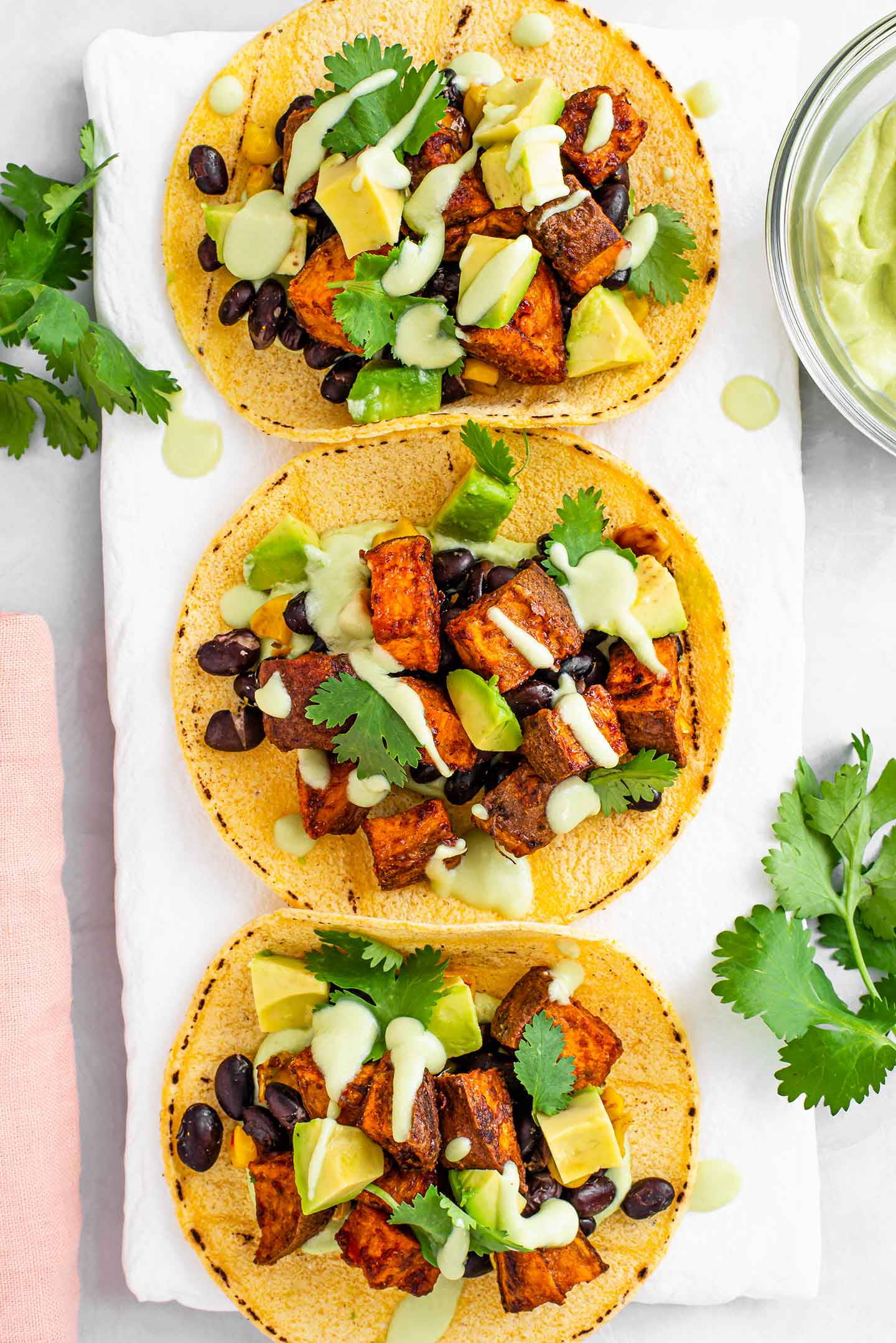 Top down view of three corn tortillas on a white tray filled with sweet potato, black beans, corn, diced avocado and drizzled with a light green avocado crema. Fresh cilantro garnishes.