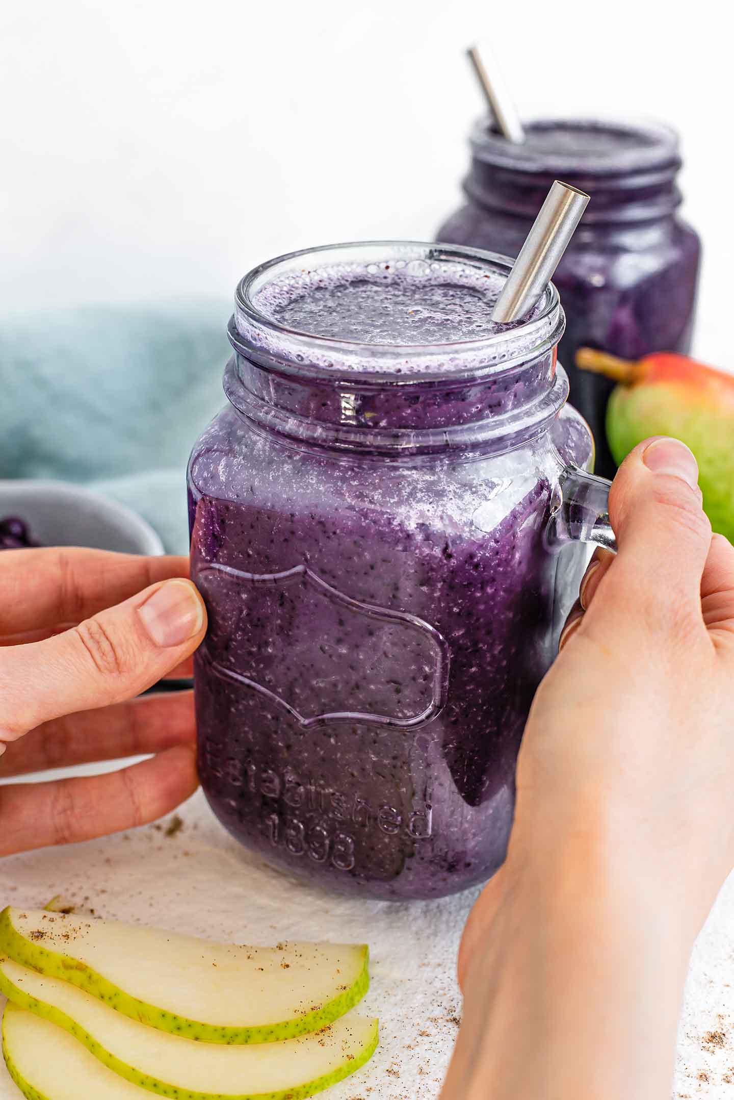 Side view of two hands holding a blueberry pear smoothie. Another smoothie is in the background and pear slices, sprinkled with cardamom are on a white tray.