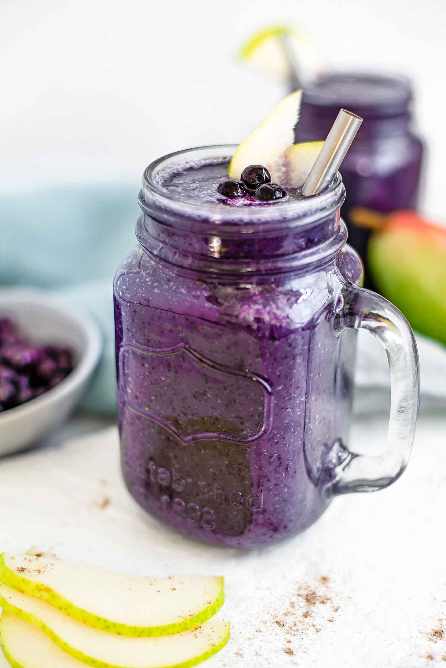 Side view of a deep purple blueberry pear smoothie in a glass jar with a handle. Slices of pear and fresh blueberries rest on top of the smoothie with a stainless steel straw.