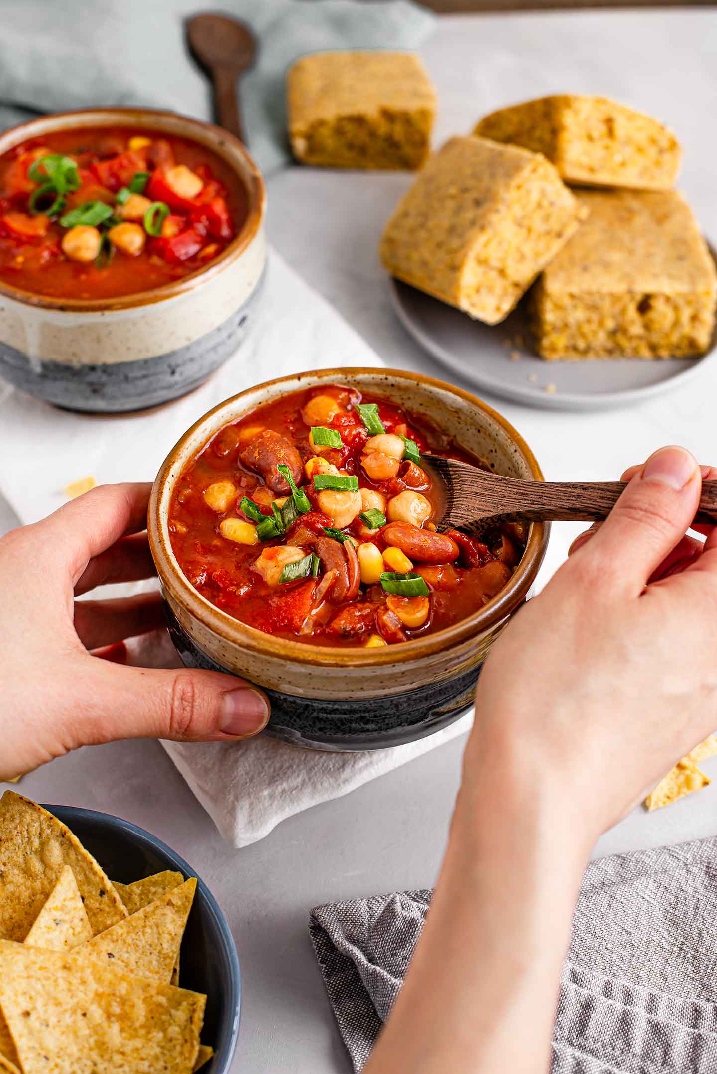 Side view of a hand using a wooden spoon to scoop easy chipotle chilli from a full, comforting bowl. Cornbread, corn chips, and another bowl of chilli are in the background.