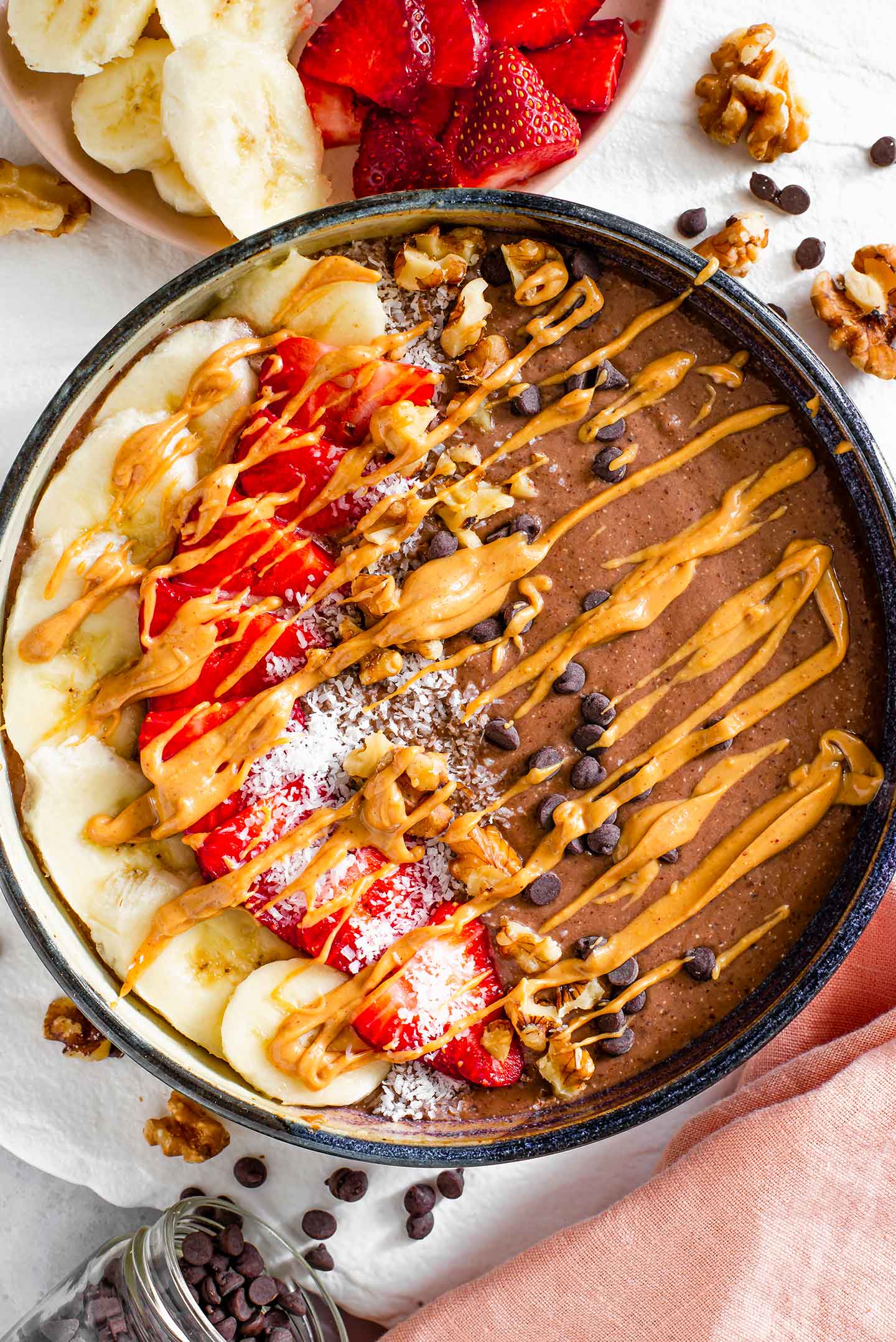Top down view of a chocolate smoothie bowl topped with banana and strawberry slices, coconut flakes, crushed walnuts, chocolate chips, and drizzled with peanut butter.