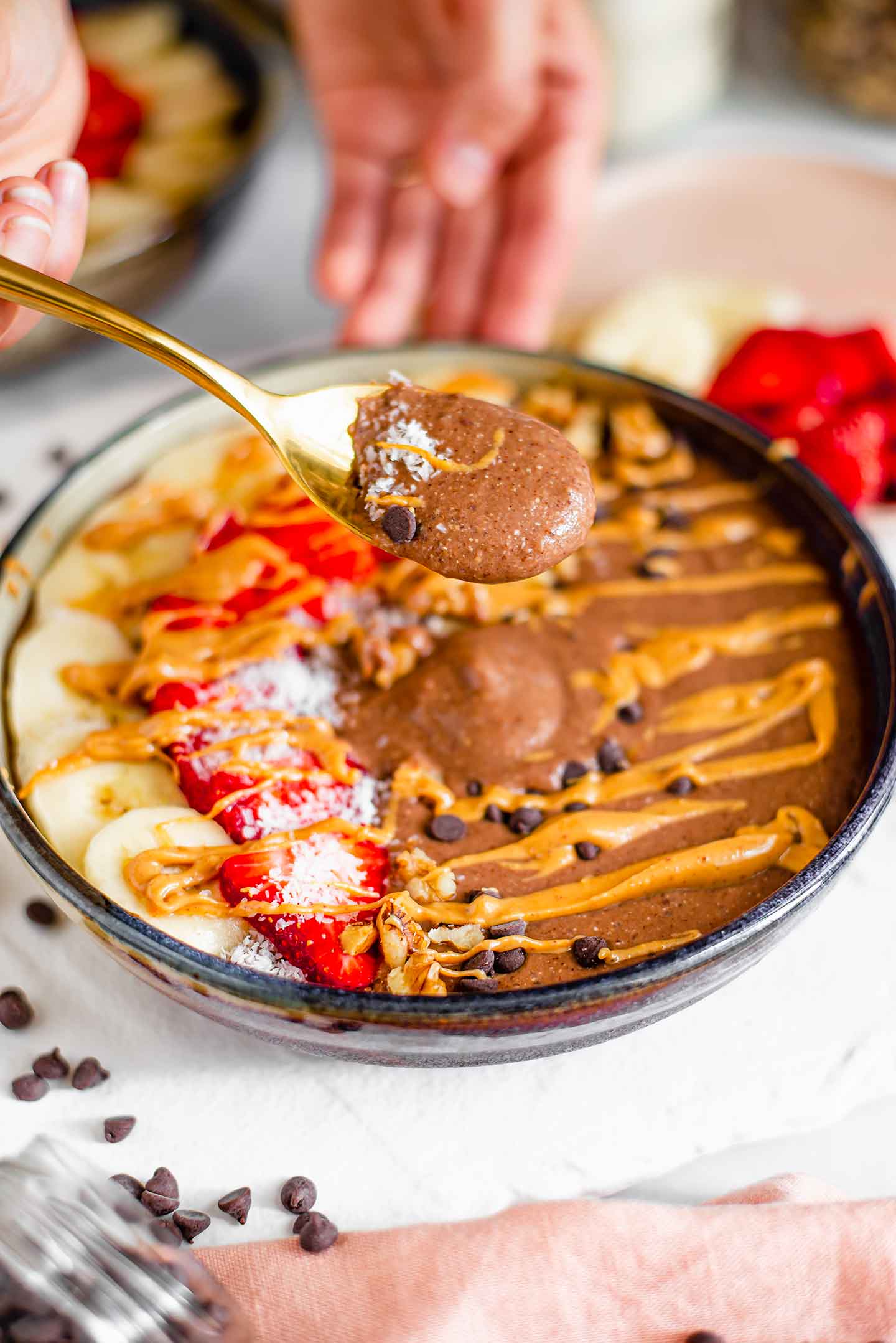 Side view of a spoon lifting thick chocolate smoothie from the decorated smoothie bowl drizzled with peanut butter. 