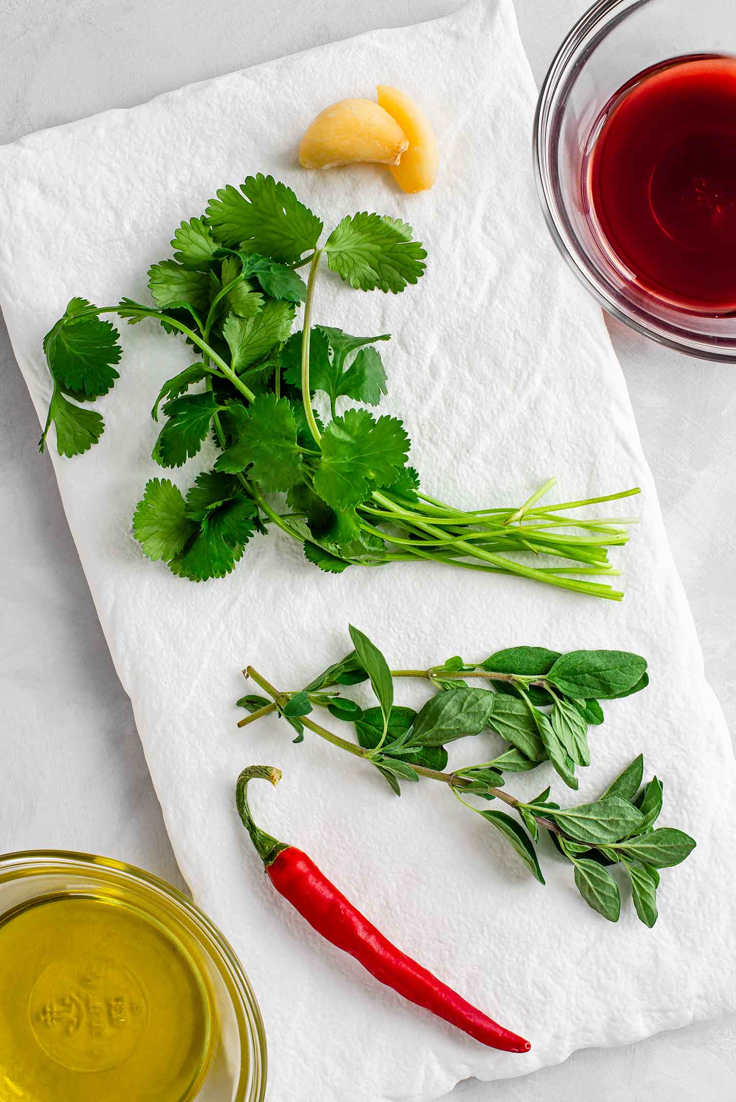 Top down view of fresh cilantro, oregano, garlic, and a chilli pepper on a white tray. Red wine vinegar and olive oil fill small bowls to the side.