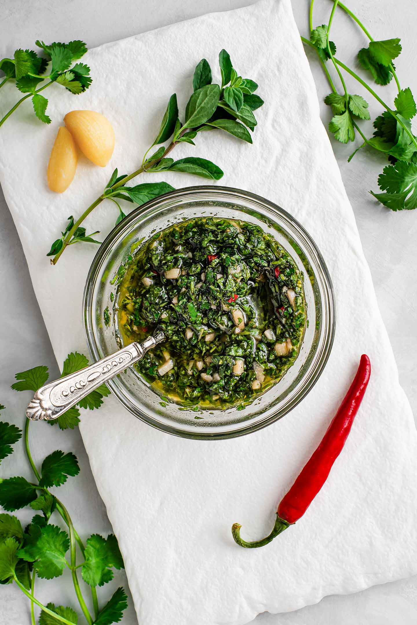 Top down view of cilantro oregano chimichurri sauce in a small dish. A red chilli pepper, garlic, cilantro, and oregano surround the sauce.