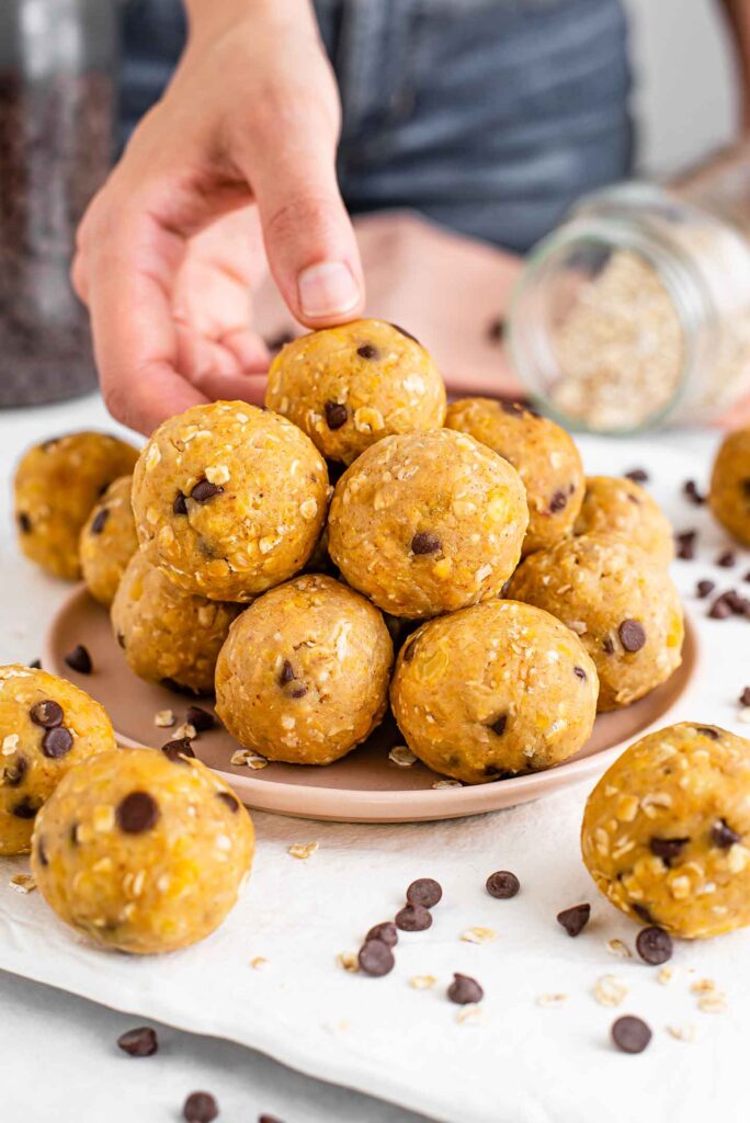 Chickpea peanut butter energy balls are stacked high on a plate. A hand grabs the top ball from the stack and chocolate chips are scattered.