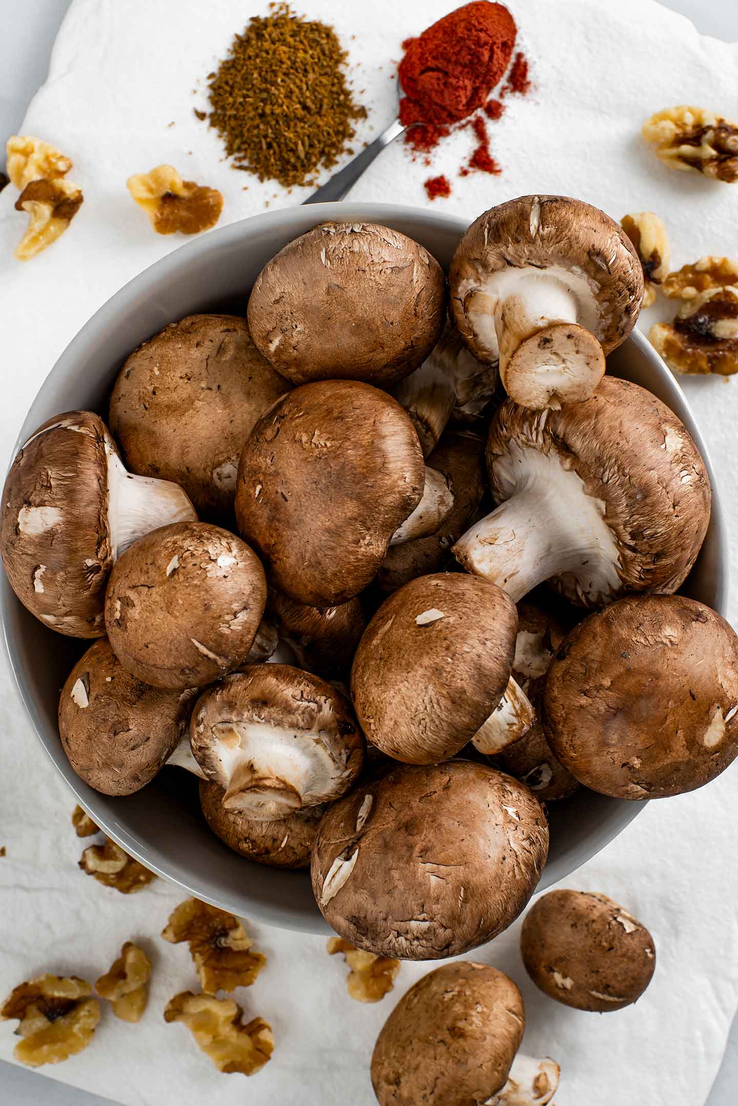 Top down view of cremini mushrooms overflowing a bowl with walnuts, cumin, and paprika on a white tray below.