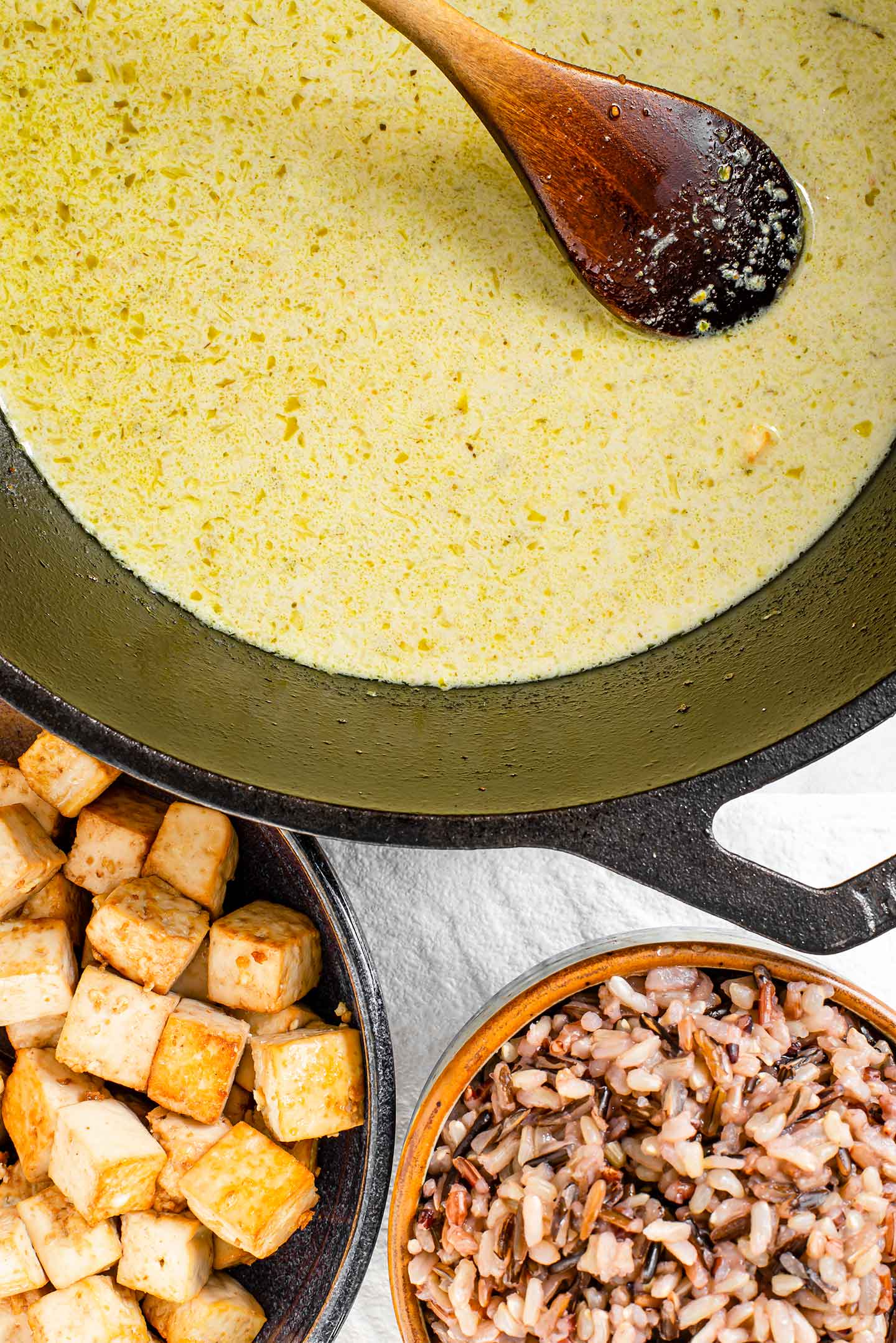 Top down view of green curry paste and coconut milk combined in a cast iron skillet with cooked wild rice and crispy tofu in separate dishes. 