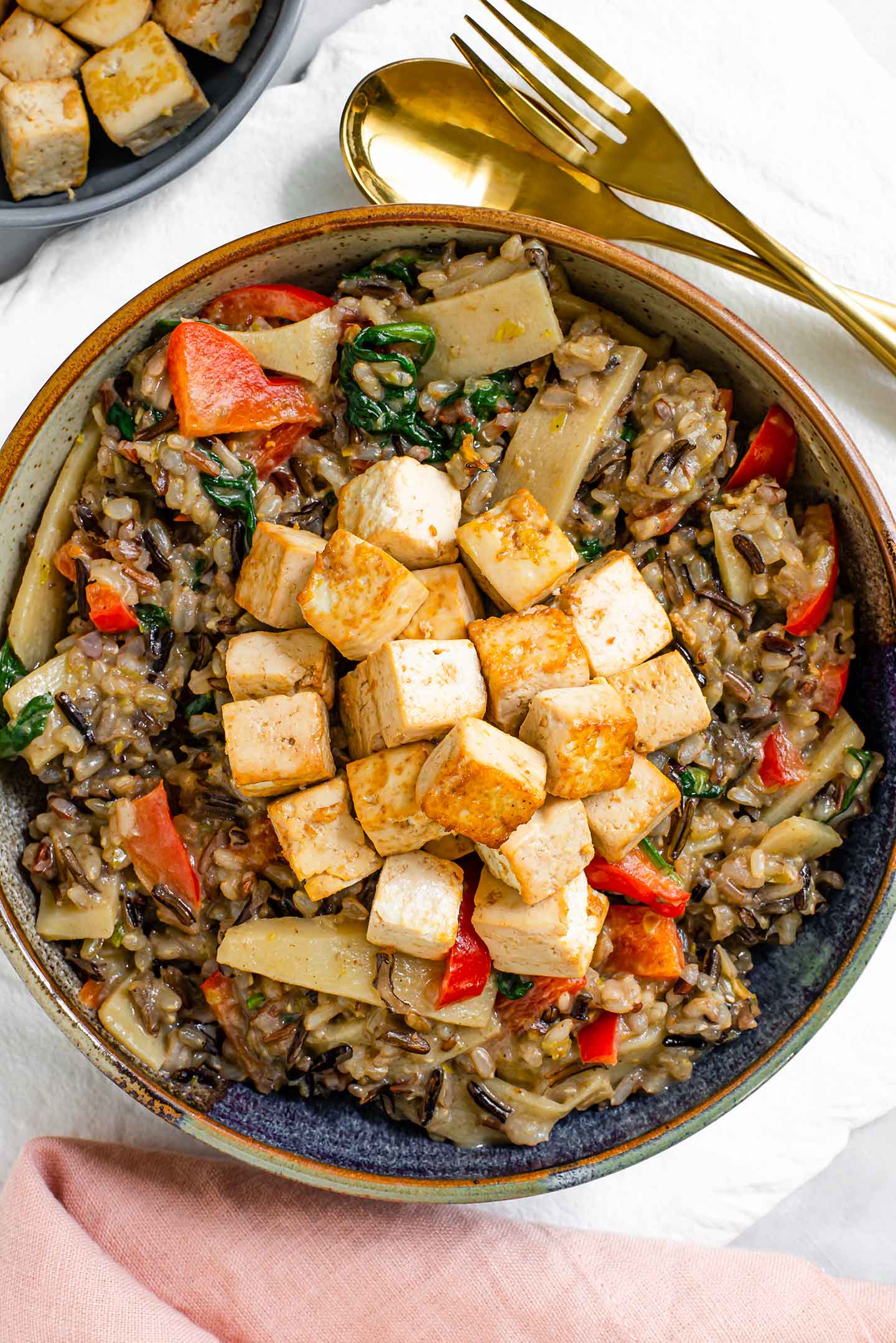 Top down view of green curry fried rice filling a shallow bowl. Wild rice mixed with bamboo shoots, red bell pepper, and spinach is topped with crispy tofu.