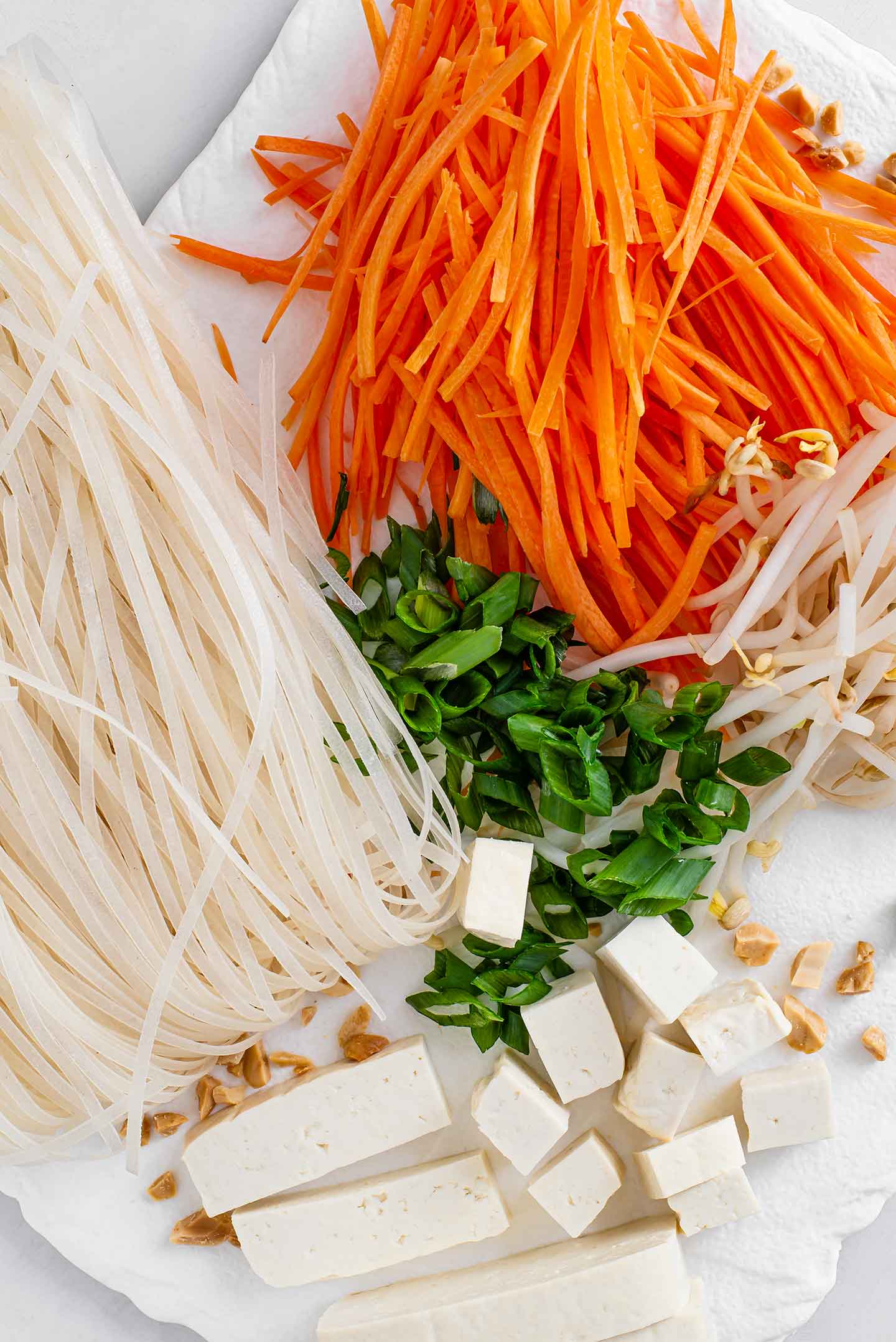 Top down view of julienned carrots, dry rice noodles, sliced green onion, bean sprouts, and cubed tofu.