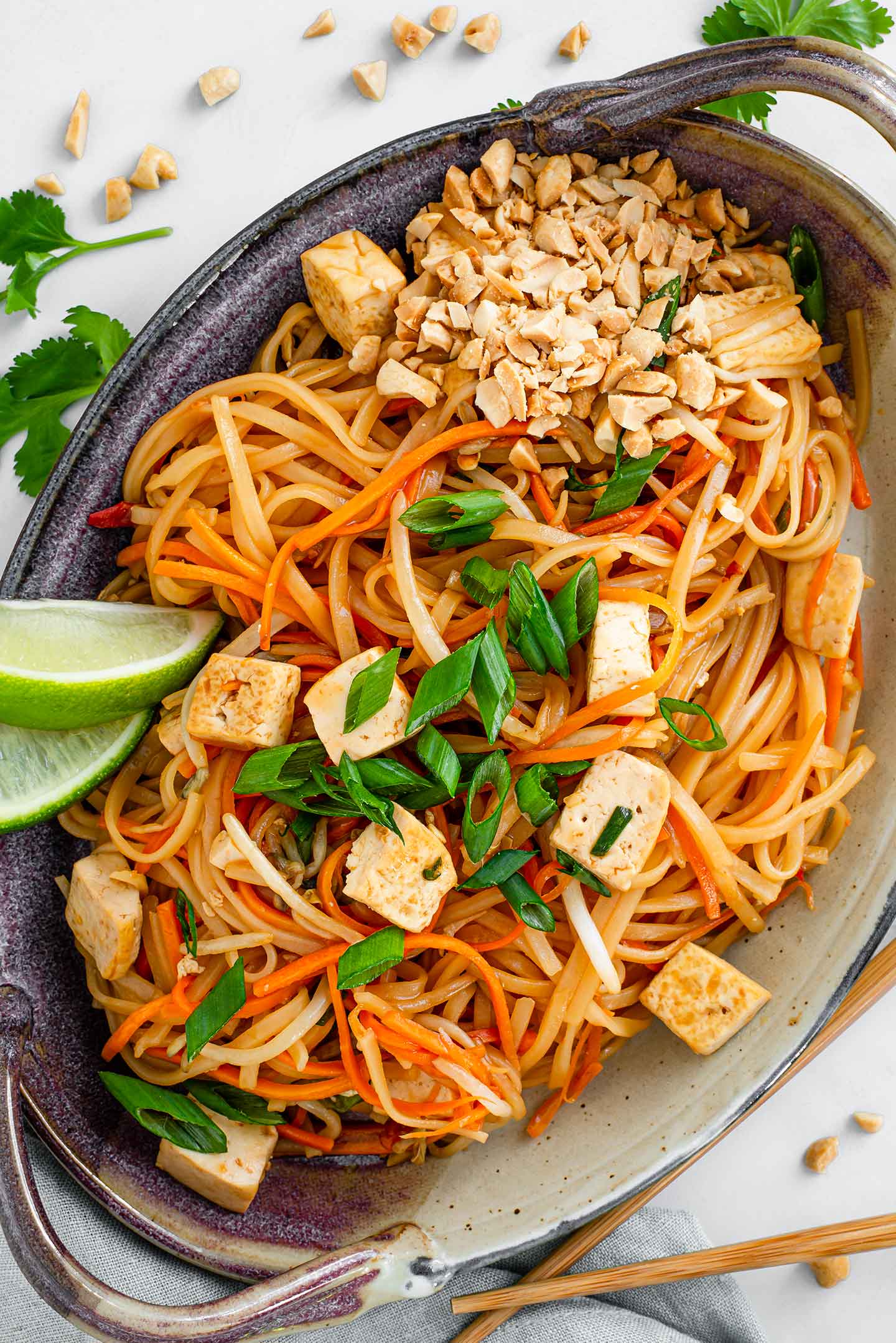 Top down view of homemade pad thai in a dish with green onion, lime wedges, and crushed peanuts for garnish.