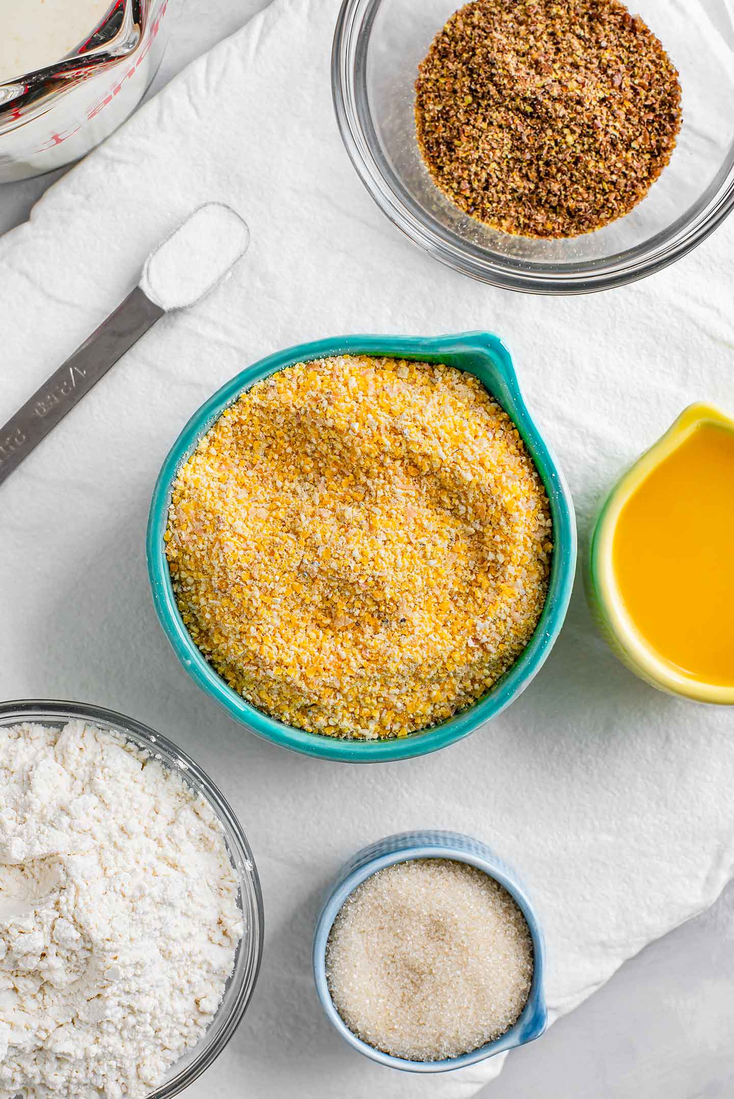 Top down view of coarse yellow cornmeal, white all purpose flour, flaxseed meal, cane sugar, melted butter and baking soda displayed on a tray. Buttermilk fills a glass measuring cup.