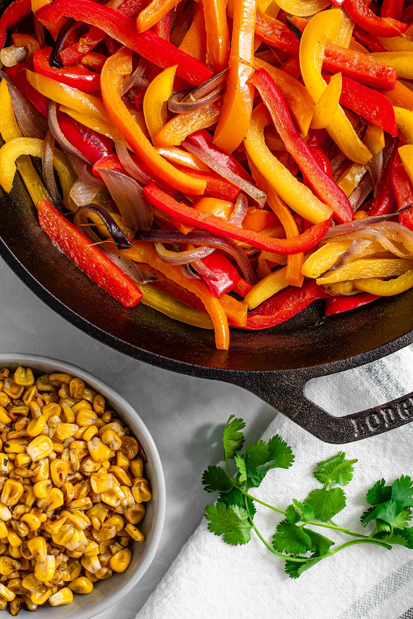 Top down view of skillet charred corn in a small dish and grilled red onion and bell peppers in a cast-iron skillet.
