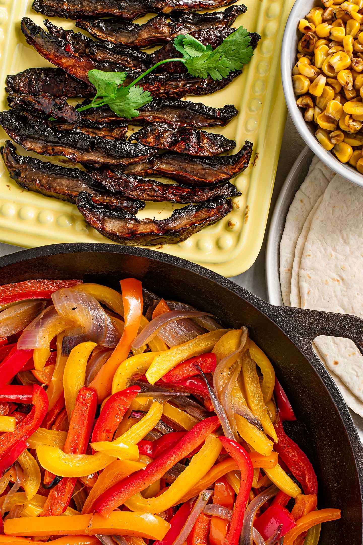 Top down view of spicy and sweet mushroom strips in a tray with grilled peppers and onion in a cast-iron skillet, charred corn in another bowl and tortillas in a plate.