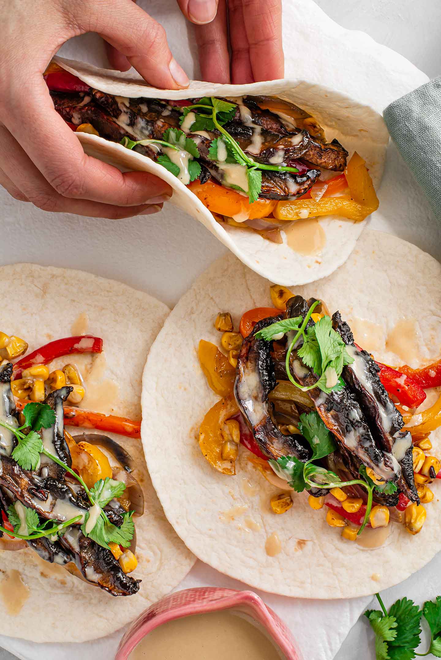 Top down view of hands picking up a fajita. Two other spicy and sweet sesame fajitas with red onion, charred corn and bell peppers lay in a tray. The fajitas are garnished with cilantro and drizzled with miso maple tahini dressing.