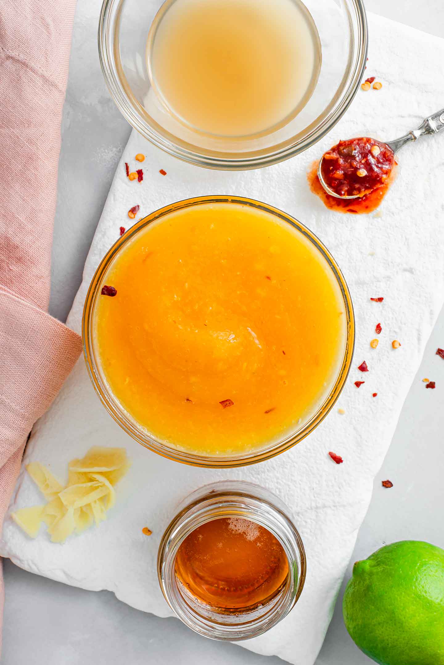 Top down view of easy mango chilli sauce in a small glass dish. Apple cider vinegar, maple syrup, fresh ginger, a lime, and chilli garlic sauce surround the dish.