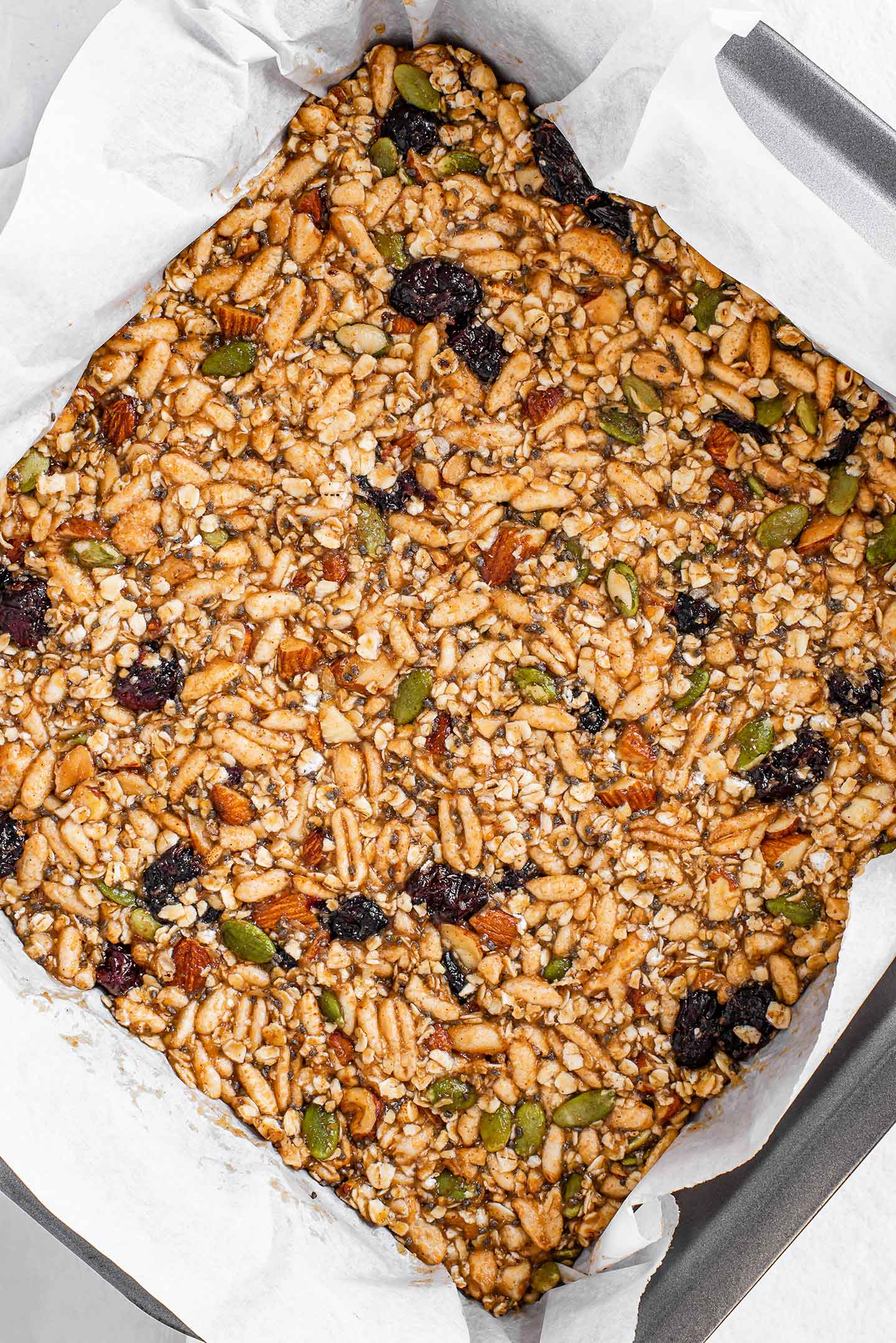 Top down view of the granola bar mixture packed into a square baking tin lined with parchment paper. The mixture looks warm and golden with dried fruit, nuts, and seeds, speckled throughout the puffed rice and oats.